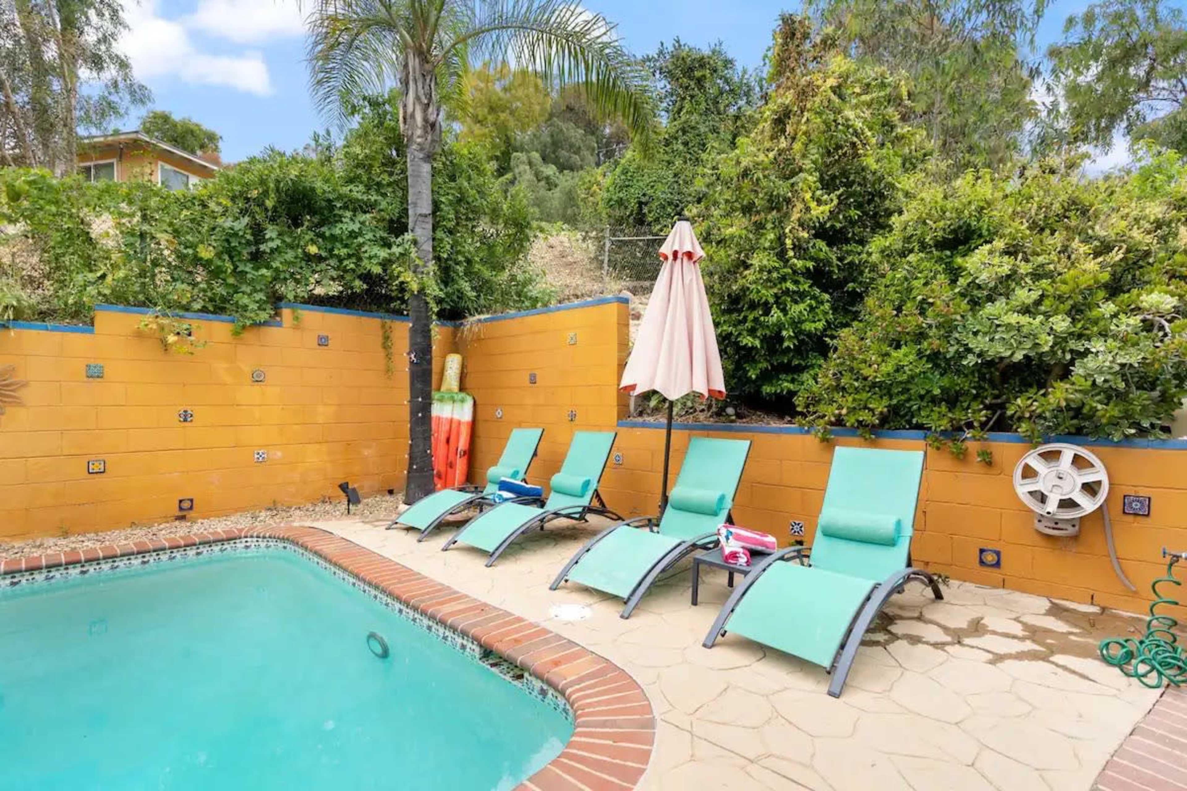 A small swimming pool is surrounded by four lounge chairs and a large umbrella, with a backdrop of lush greenery and a yellow wall.