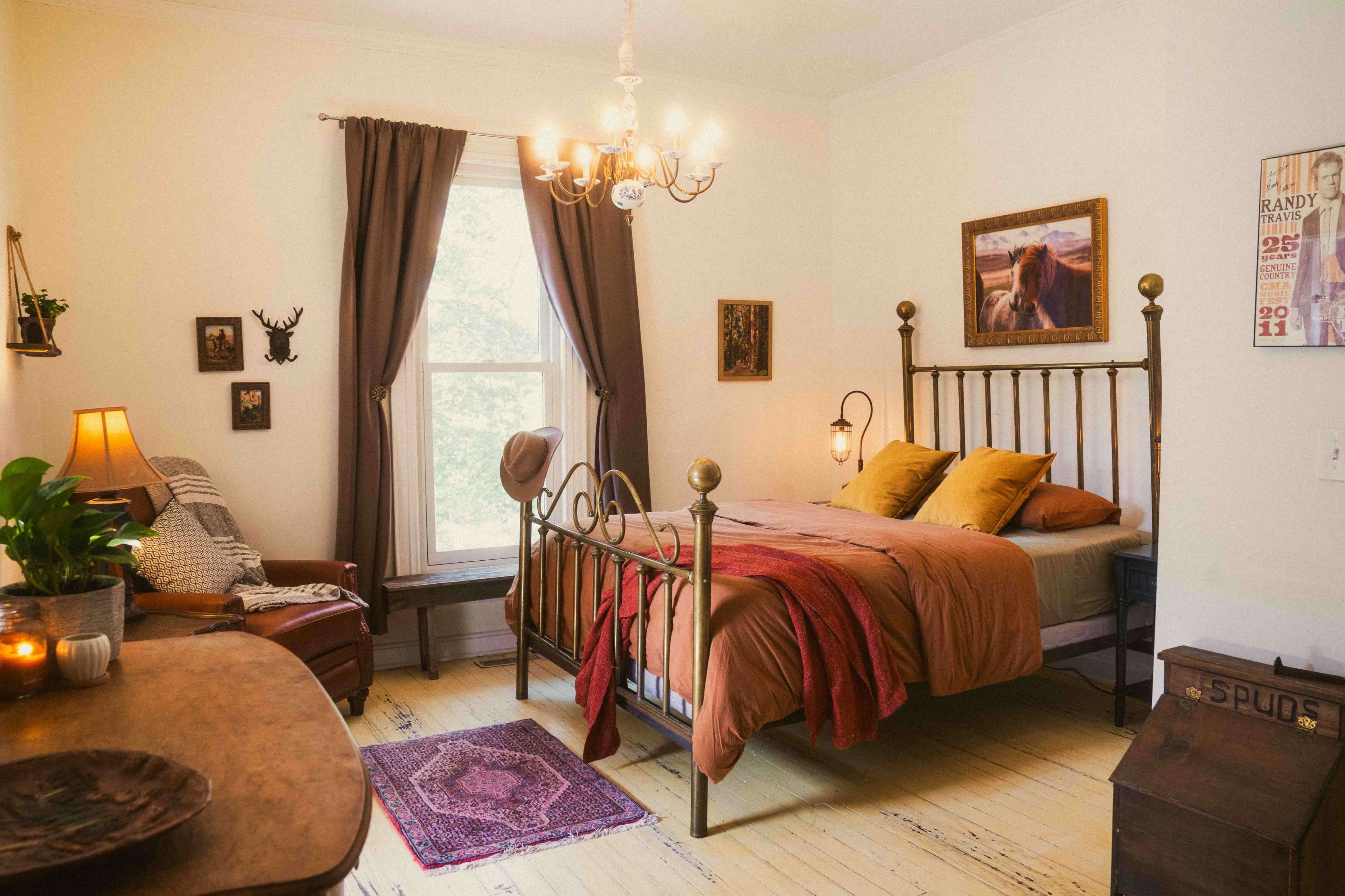 The image features a cozy bedroom with a brass bed frame, a wooden table, armchair, and warm lighting, surrounded by decorative wall art and a window allowing natural light.