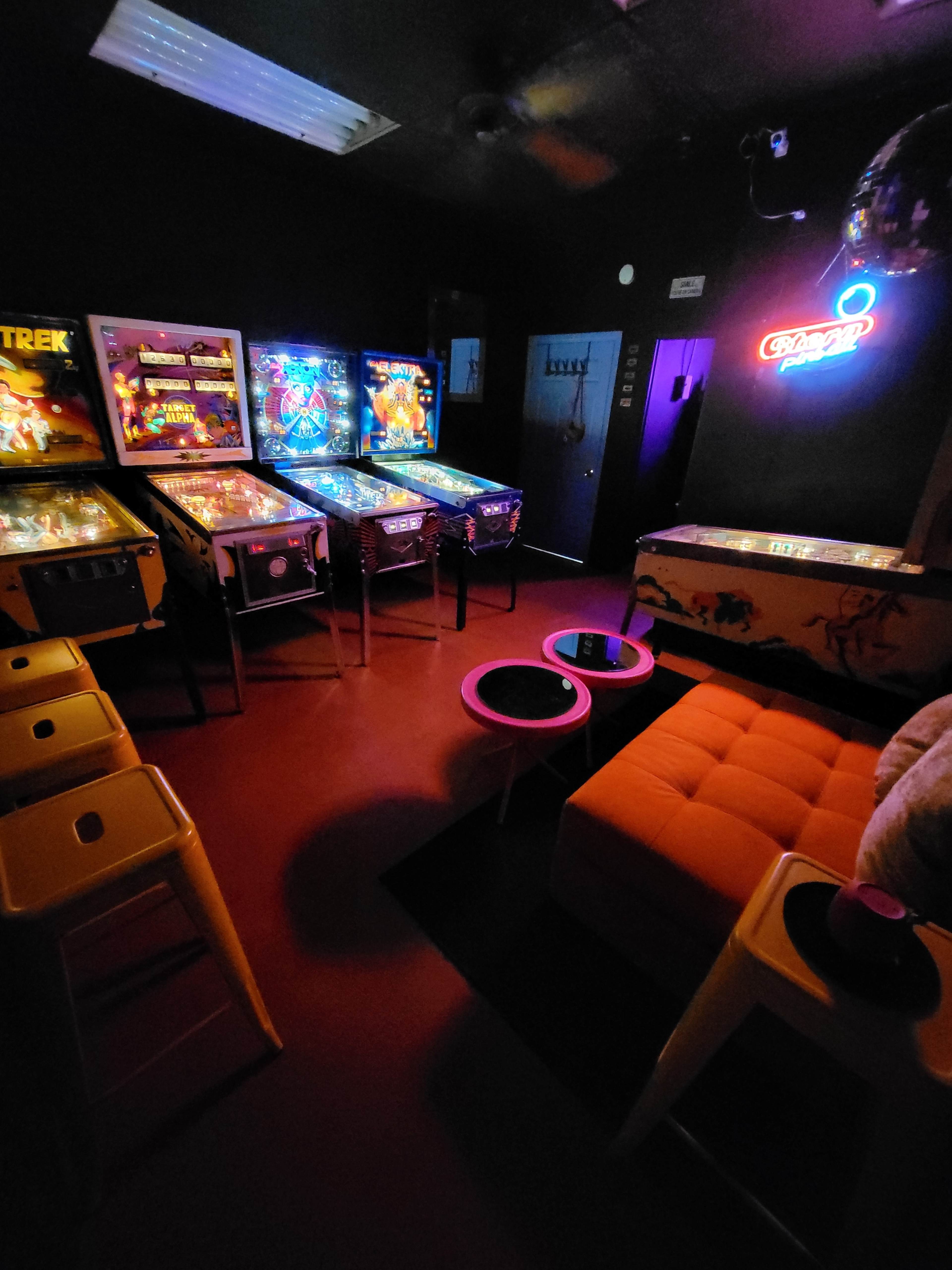 A dimly lit room featuring several pinball machines, neon lighting, and colorful furniture, including an orange couch and yellow stools.
