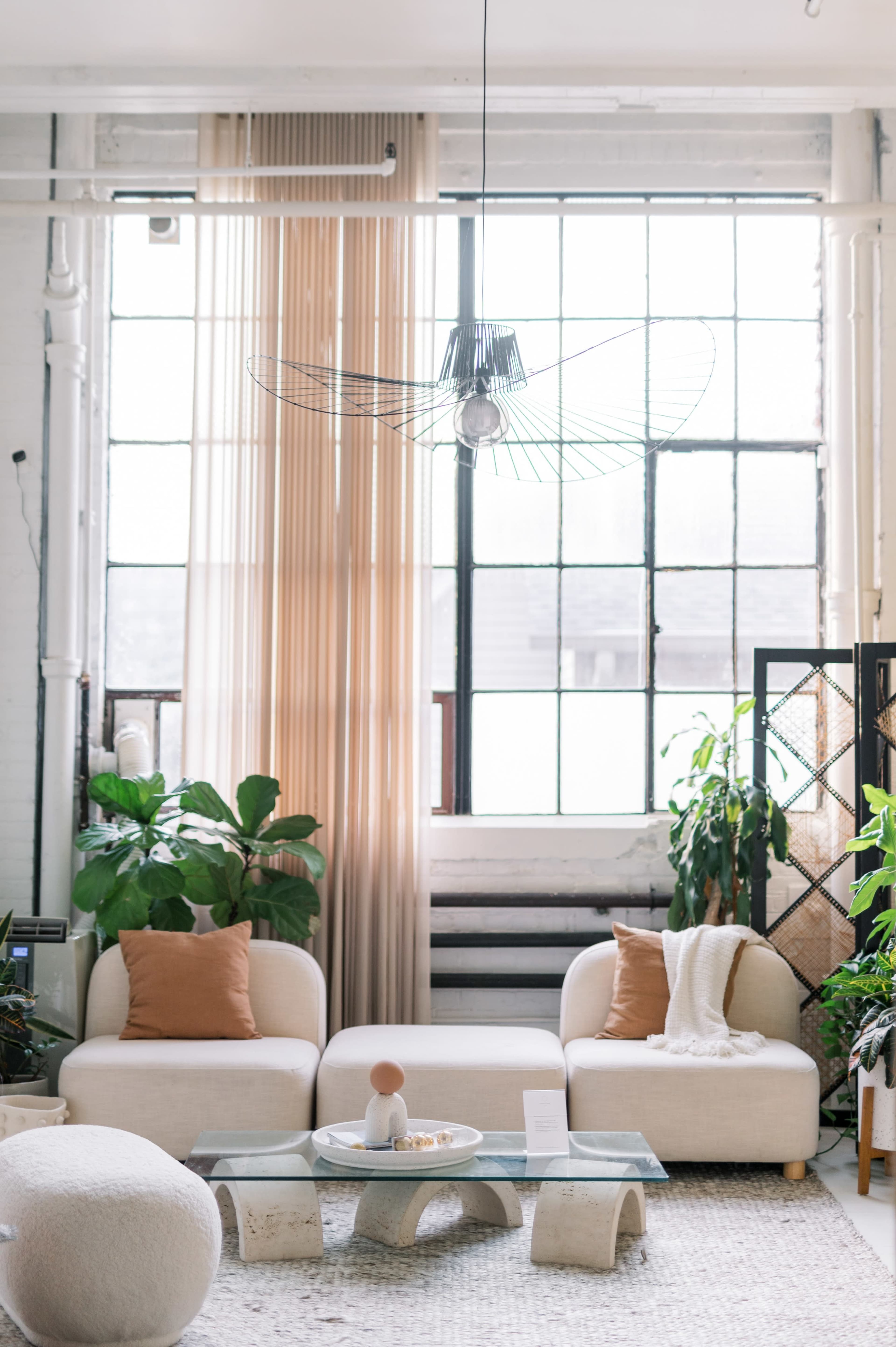 A spacious living room features two white sofas with orange cushions, a glass coffee table, and large windows draped with sheer curtains, surrounded by various indoor plants.