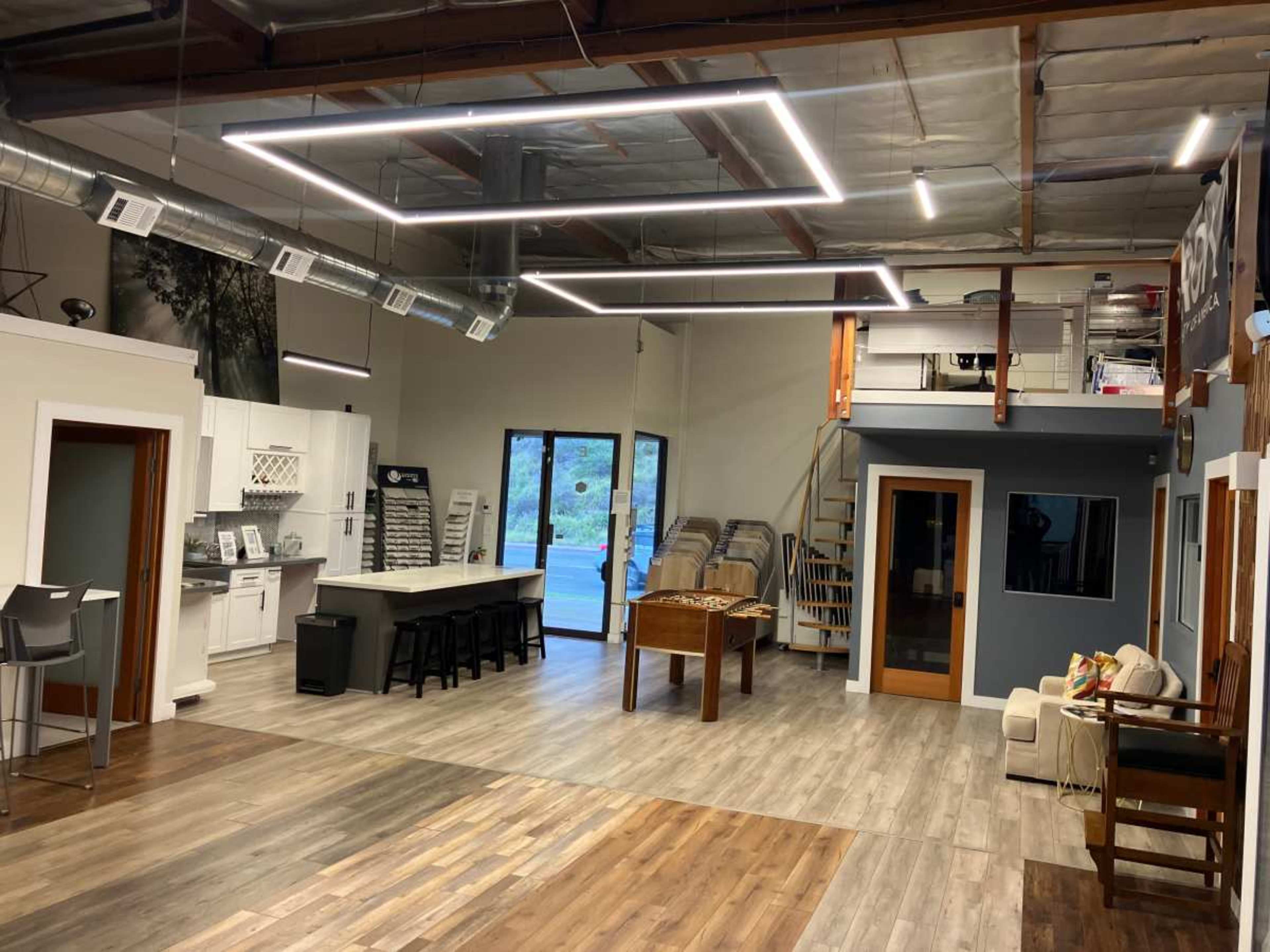 The image shows a spacious, well-lit interior with modern lighting, featuring a kitchen area, a staircase, seating, and a game table.