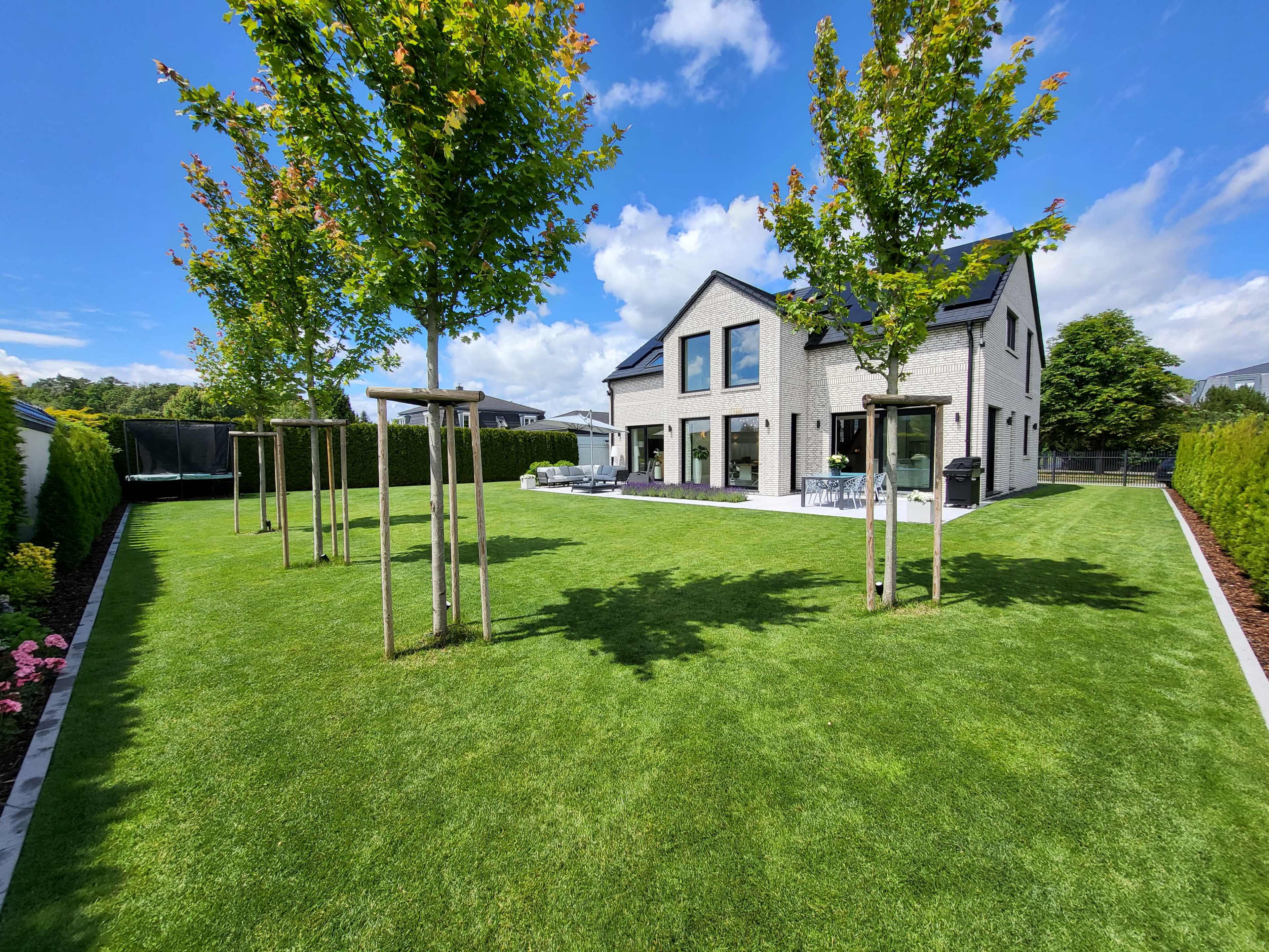 A modern house with large windows is surrounded by a well-maintained lawn and young trees, under a clear blue sky.