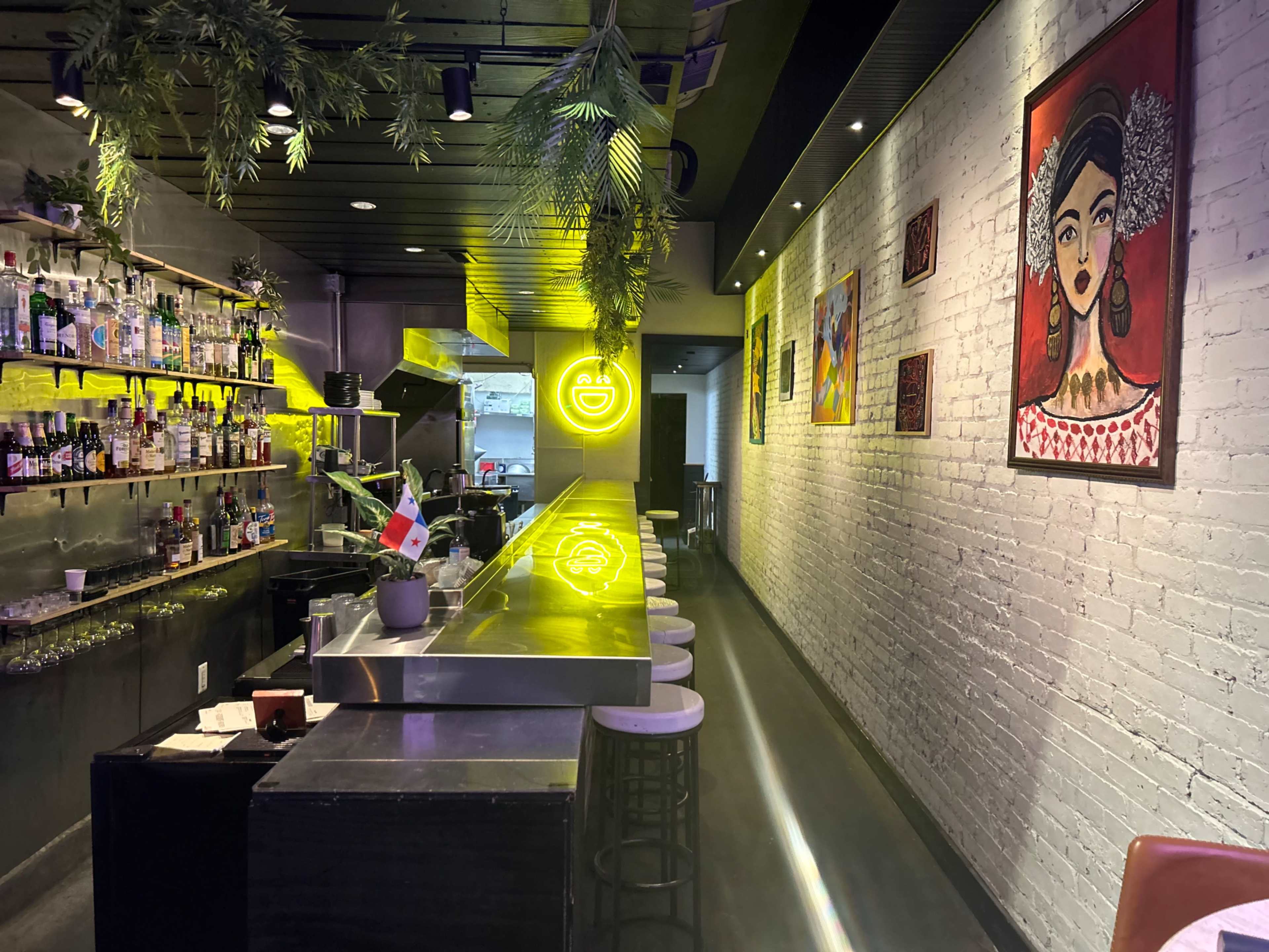 The image shows a modern bar with a neon sign, a well-stocked shelf of liquor, and vibrant artwork on the walls.