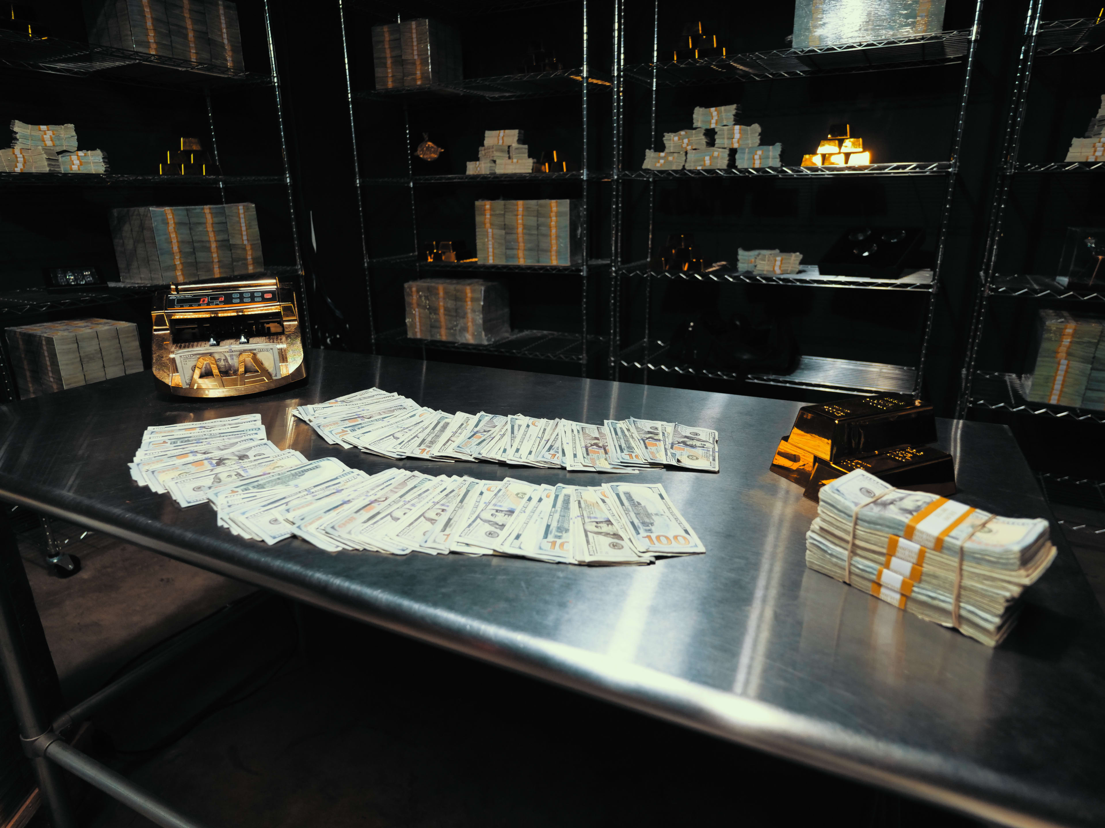 A stainless steel table displays neatly arranged stacks of cash, with additional bundles near a small vehicle and various items in the background on shelves.