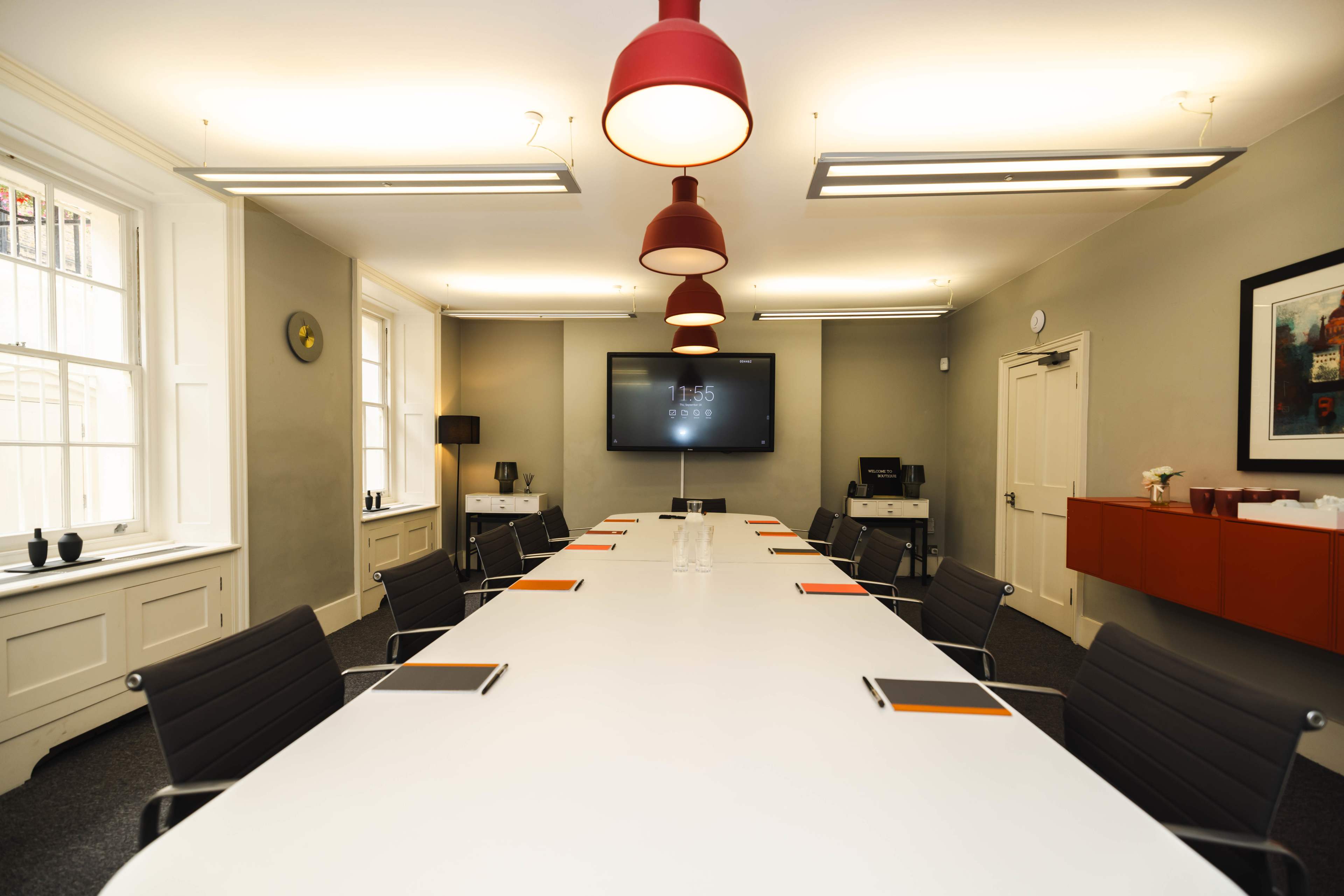A modern conference room features a long white table surrounded by black chairs, with a large screen mounted on the wall and red pendant lights overhead.
