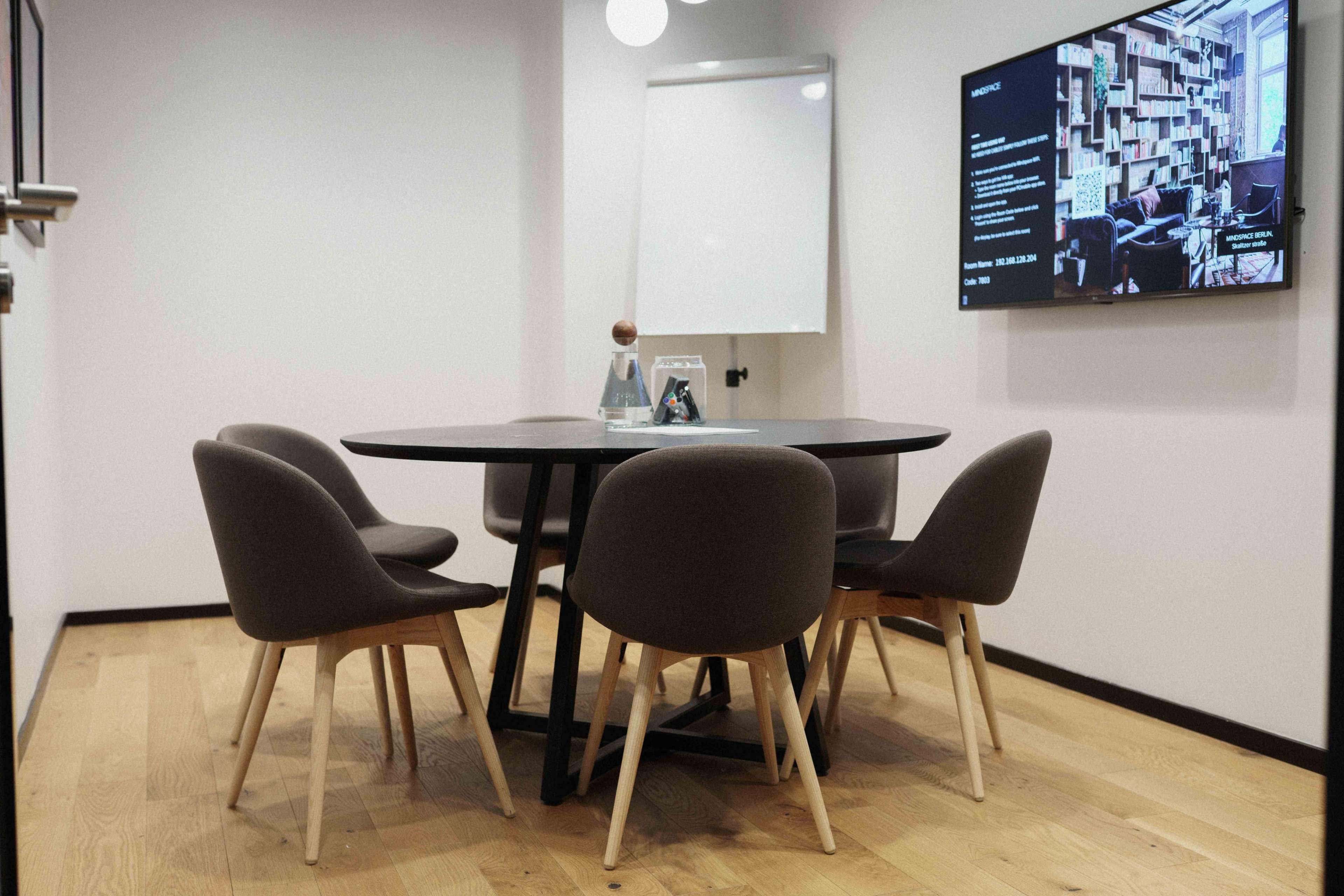 A modern conference room features a round table surrounded by five chairs, a whiteboard, and a wall-mounted display.
