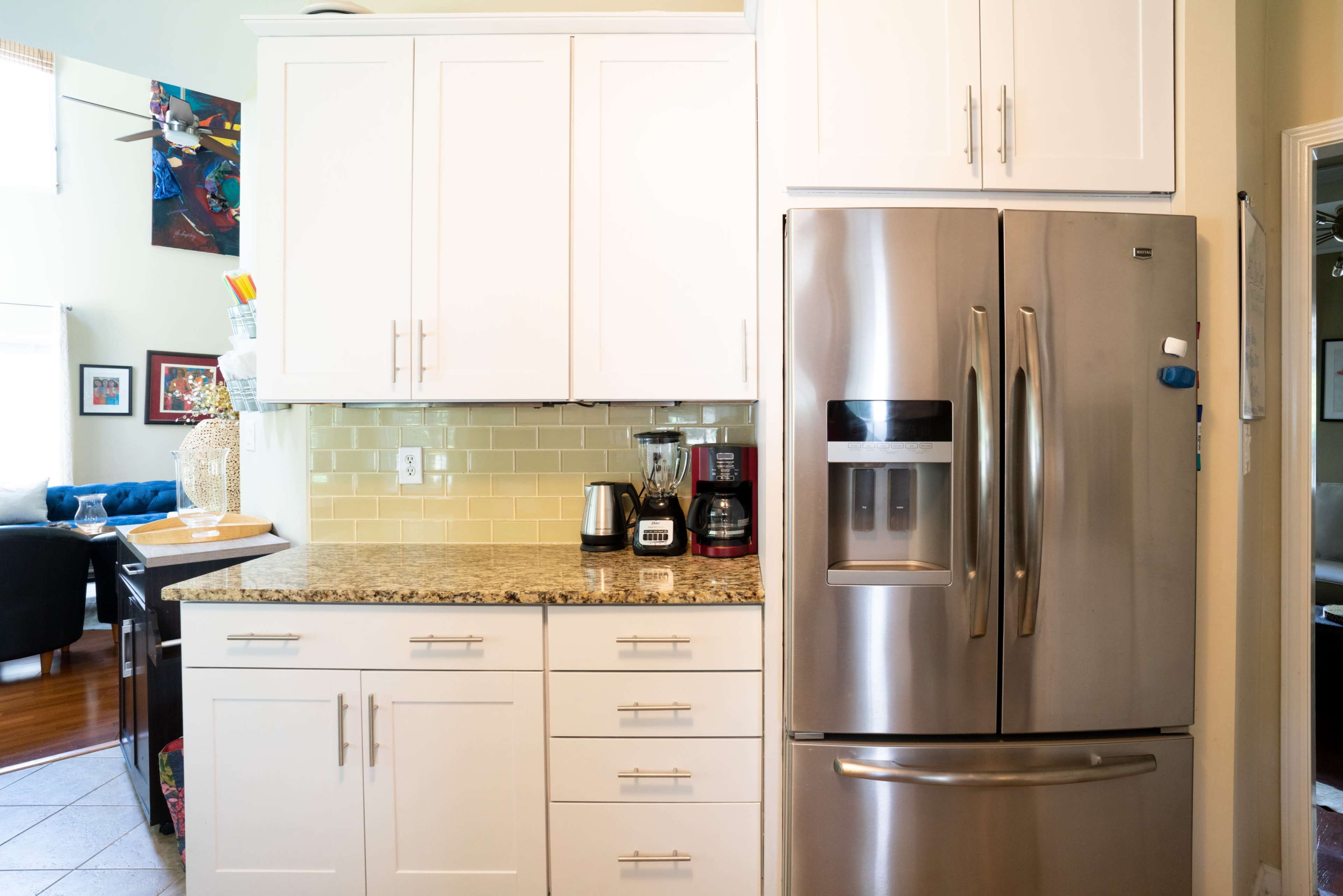 The image shows a modern kitchen featuring white cabinetry, a stainless steel refrigerator, and a granite countertop with small appliances.