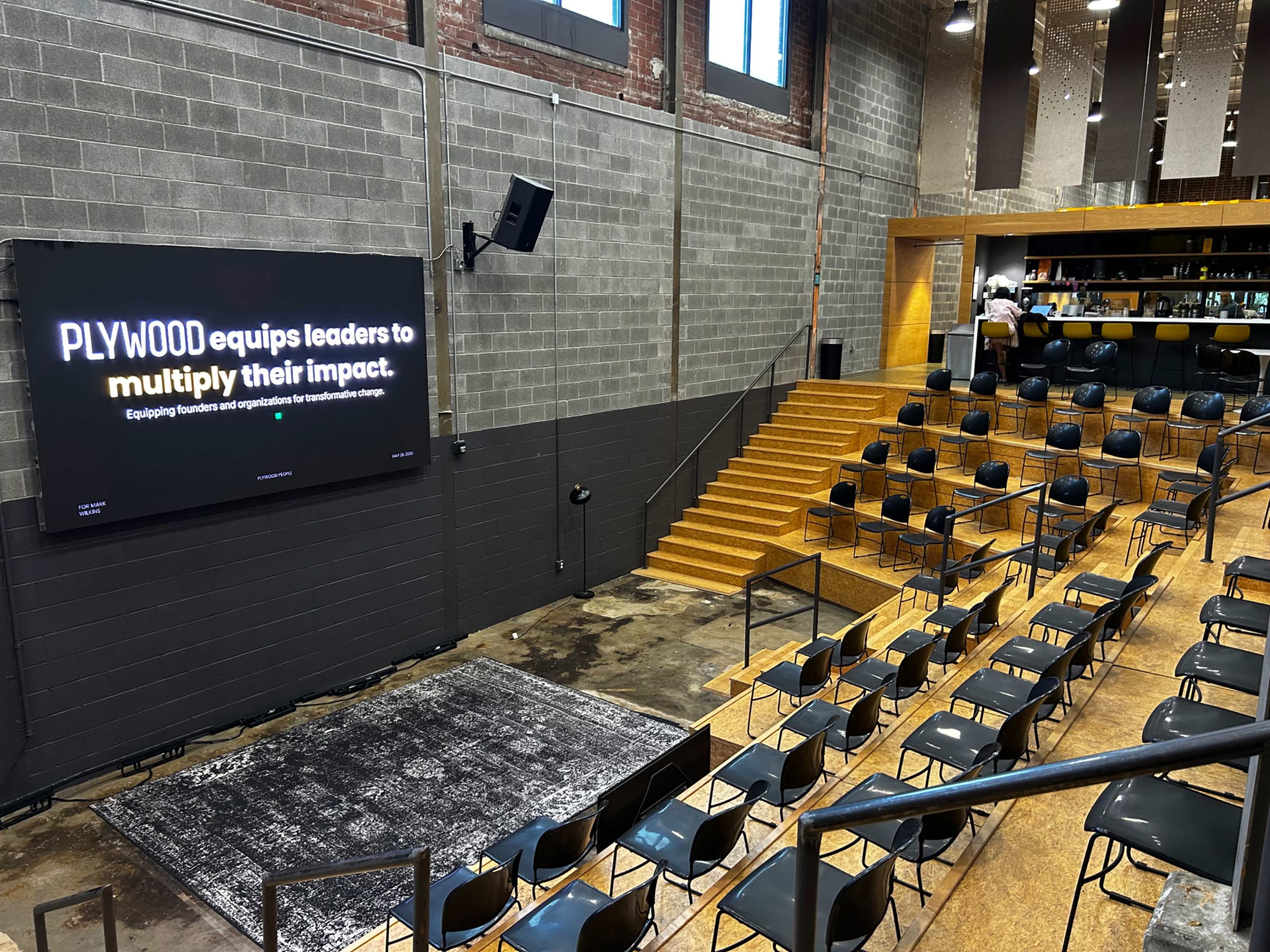 The image shows a modern indoor venue with rows of chairs arranged for an event, featuring a sign on the wall that reads "PLYWOOD equips leaders to multiply their impact."