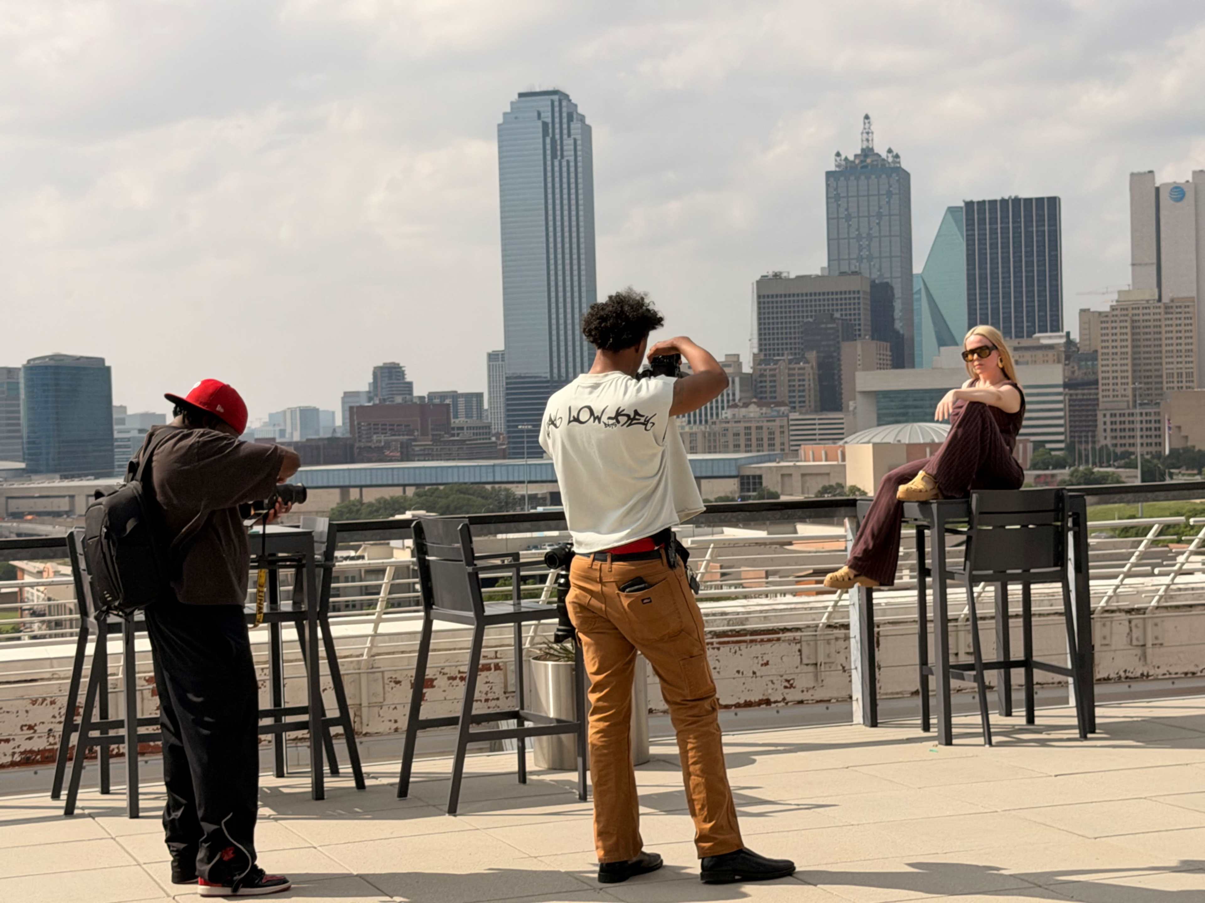 Two photographers are capturing images of a model posing on a rooftop with a city skyline in the background.