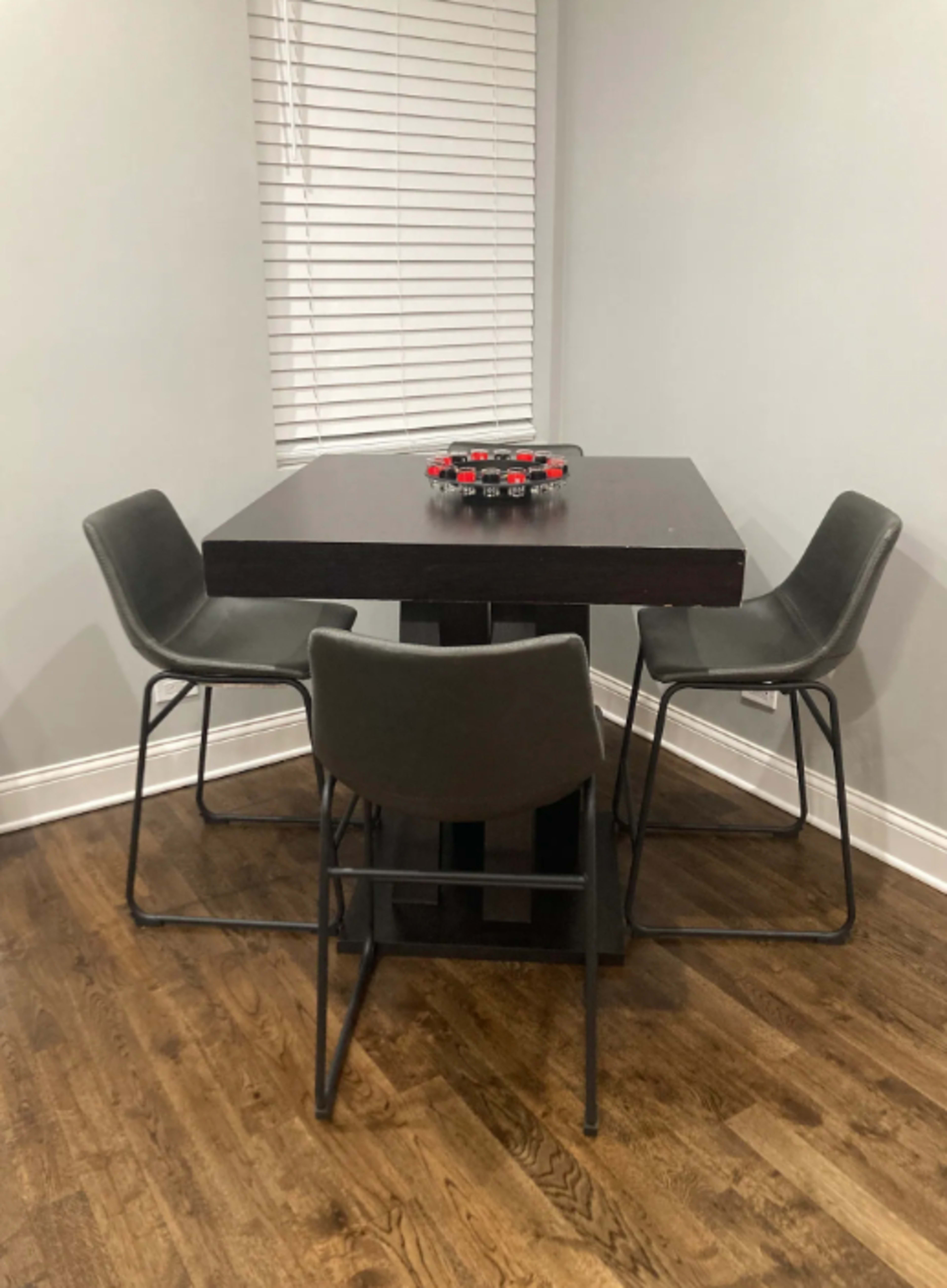 A square table with four chairs is set in a room with gray walls and a window covered by blinds.