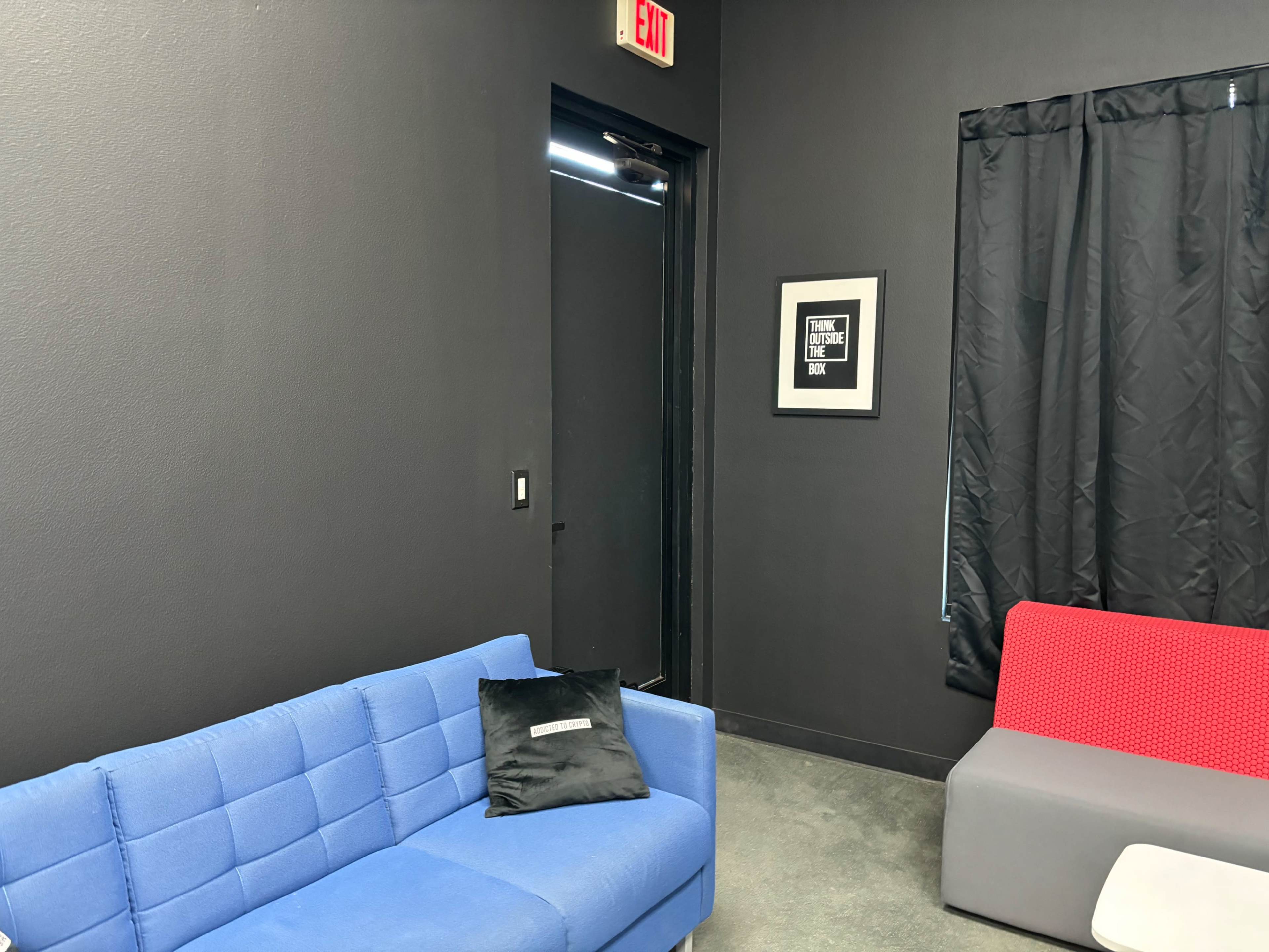 The image shows a dark-walled room with a blue sofa, a gray couch with red accents, a black curtain, and a door leading outside.