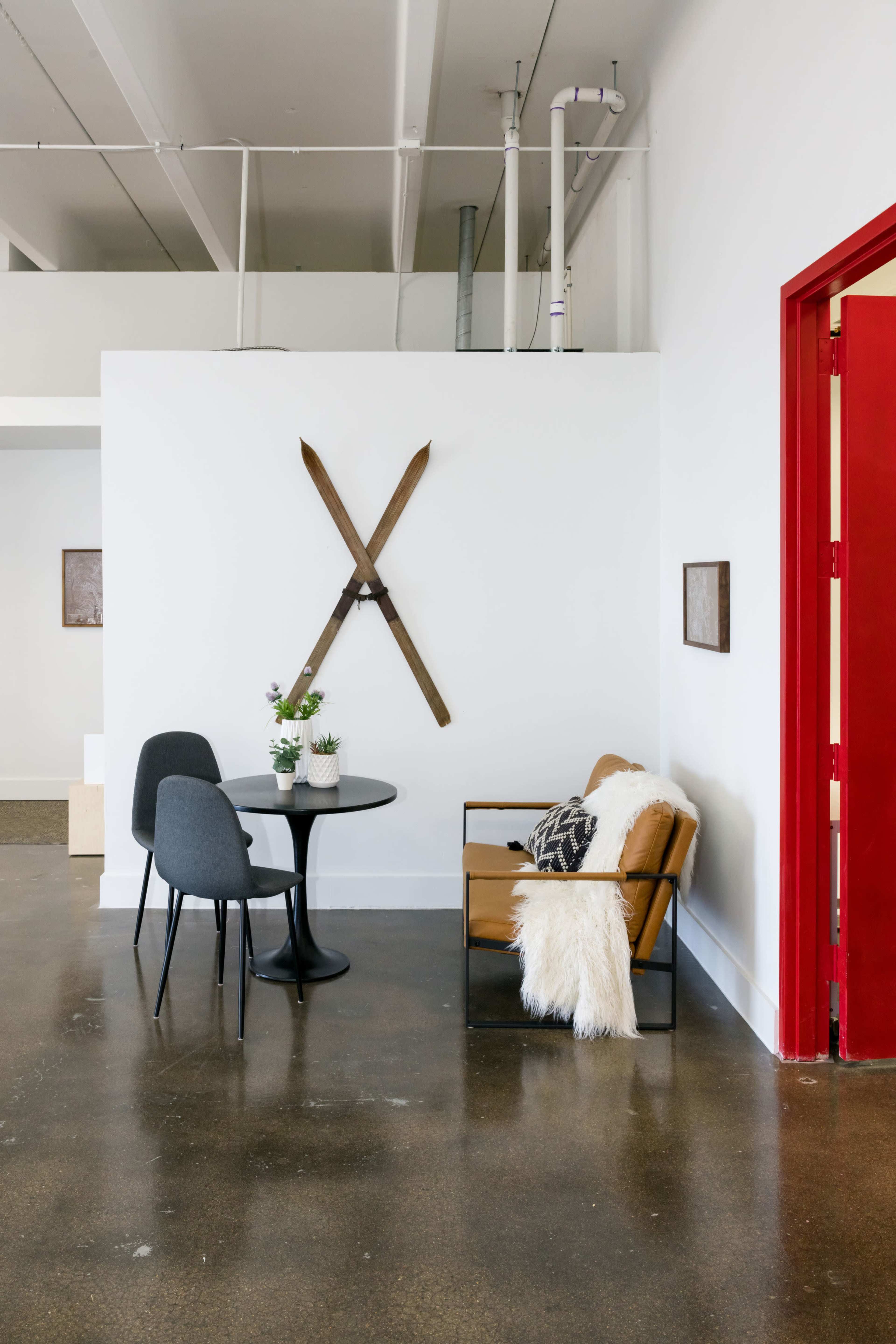 A modern interior features a round black table with two chairs, a cozy armchair with a throw, and vintage skis mounted on a white wall beside a red door.