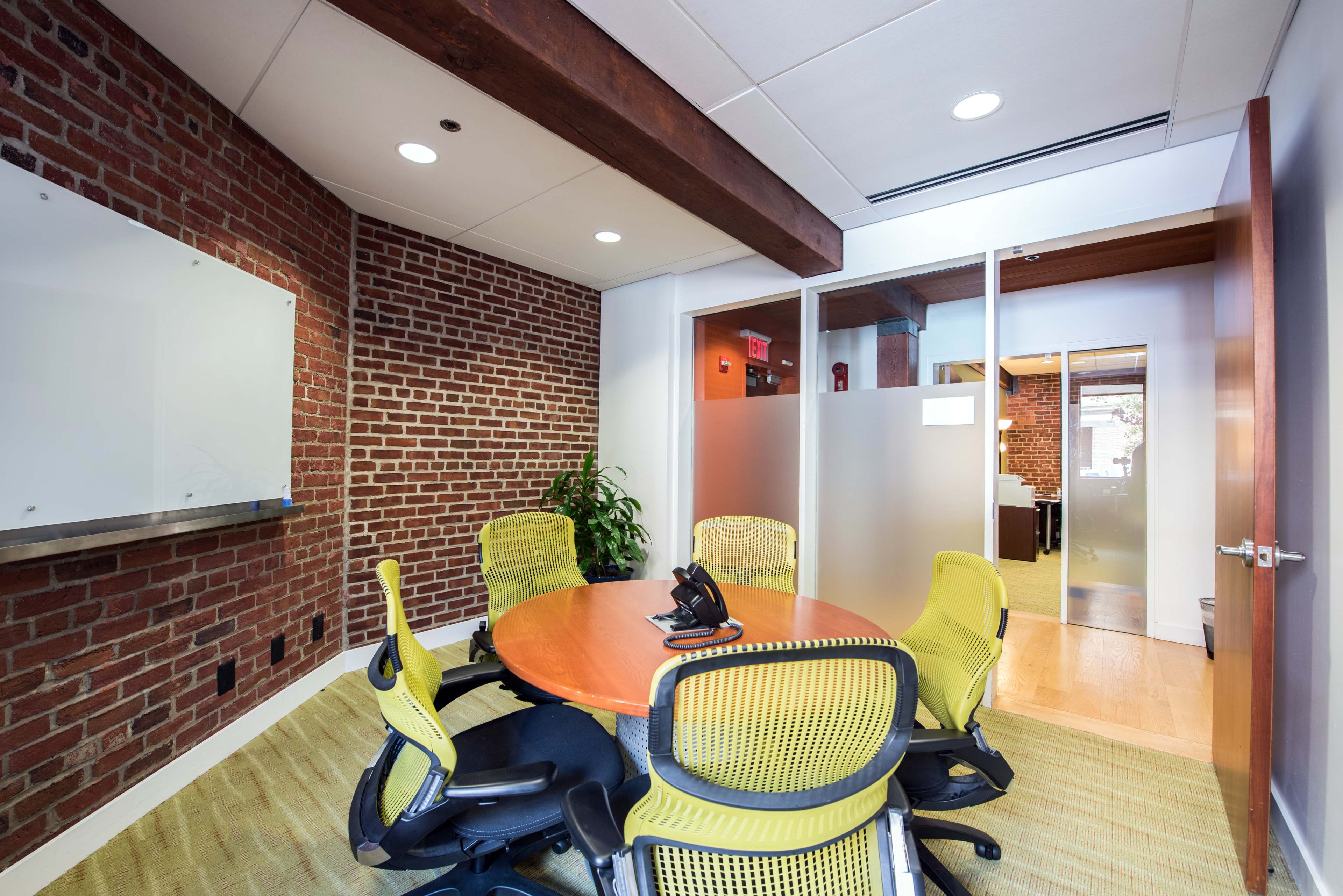 A conference room with a round wooden table surrounded by four yellow office chairs, set against a backdrop of exposed brick walls.