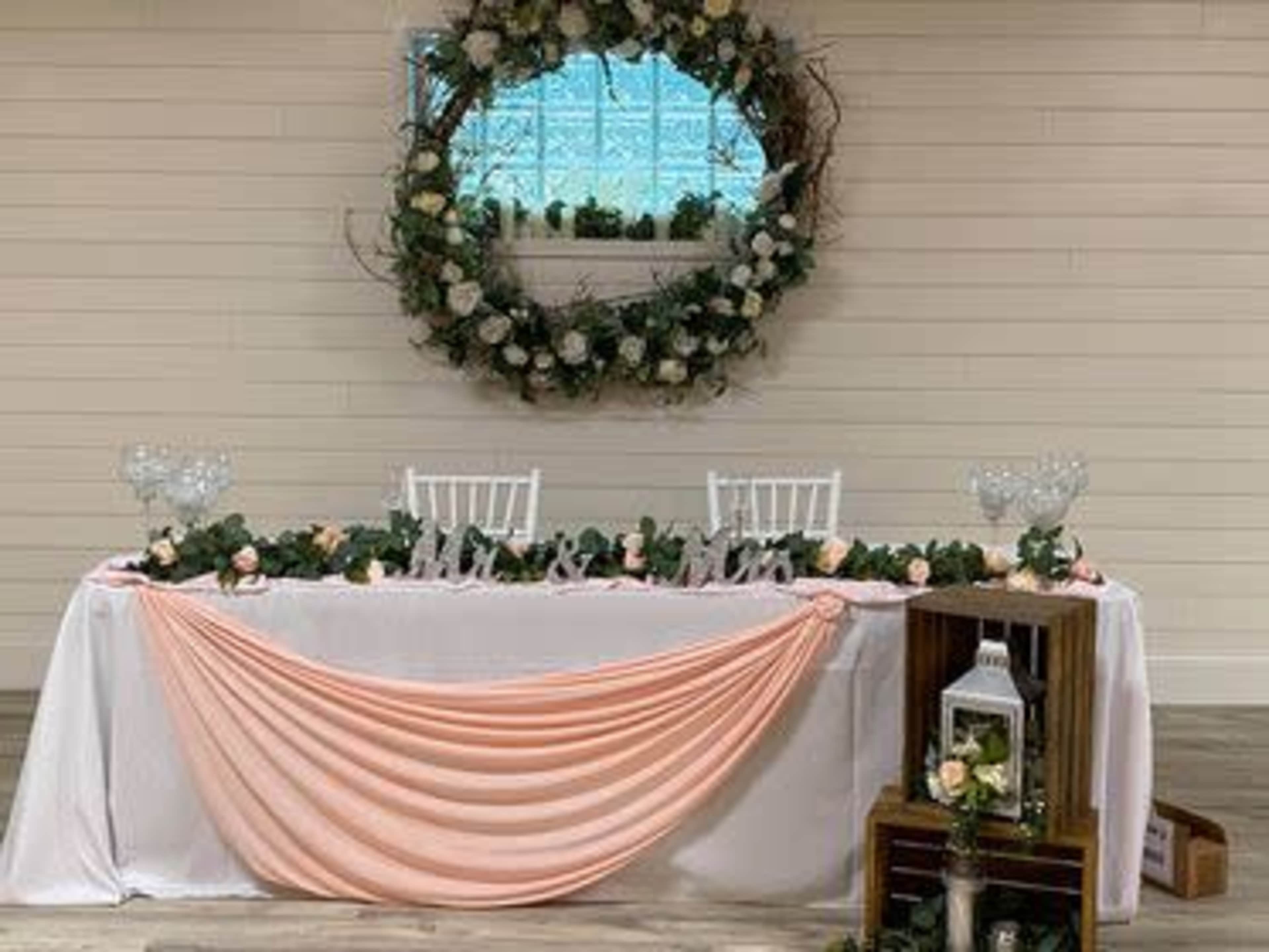 A decorated table with a white tablecloth and peach drapery is set for a wedding, featuring floral accents and a large wreath with greenery and flowers in the background.