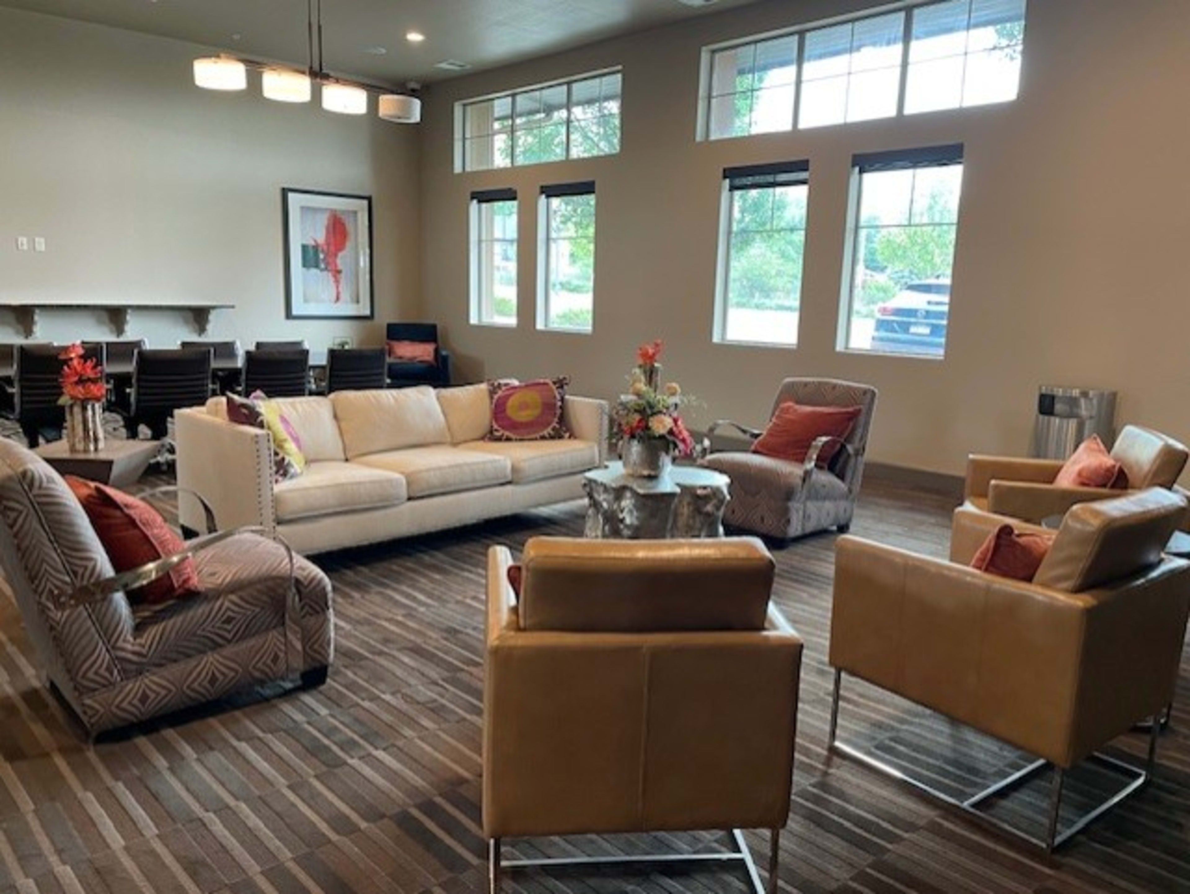 A cozy lounge area features a white sofa, two armchairs, and patterned chairs arranged around a small table, with large windows allowing natural light to enter.