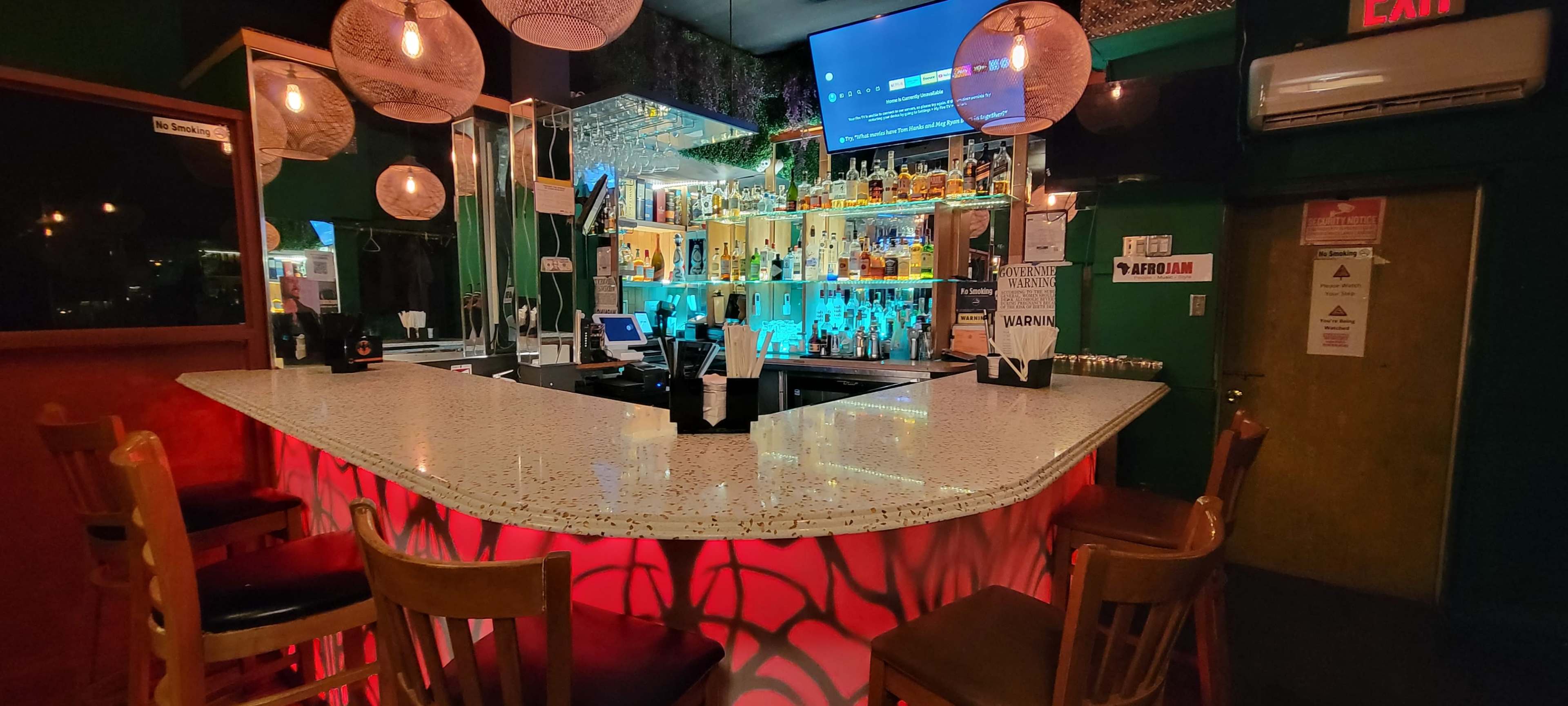 The image shows a bar with a brightly lit counter, shelves stocked with various bottles of alcohol, and wooden stools arranged around it.