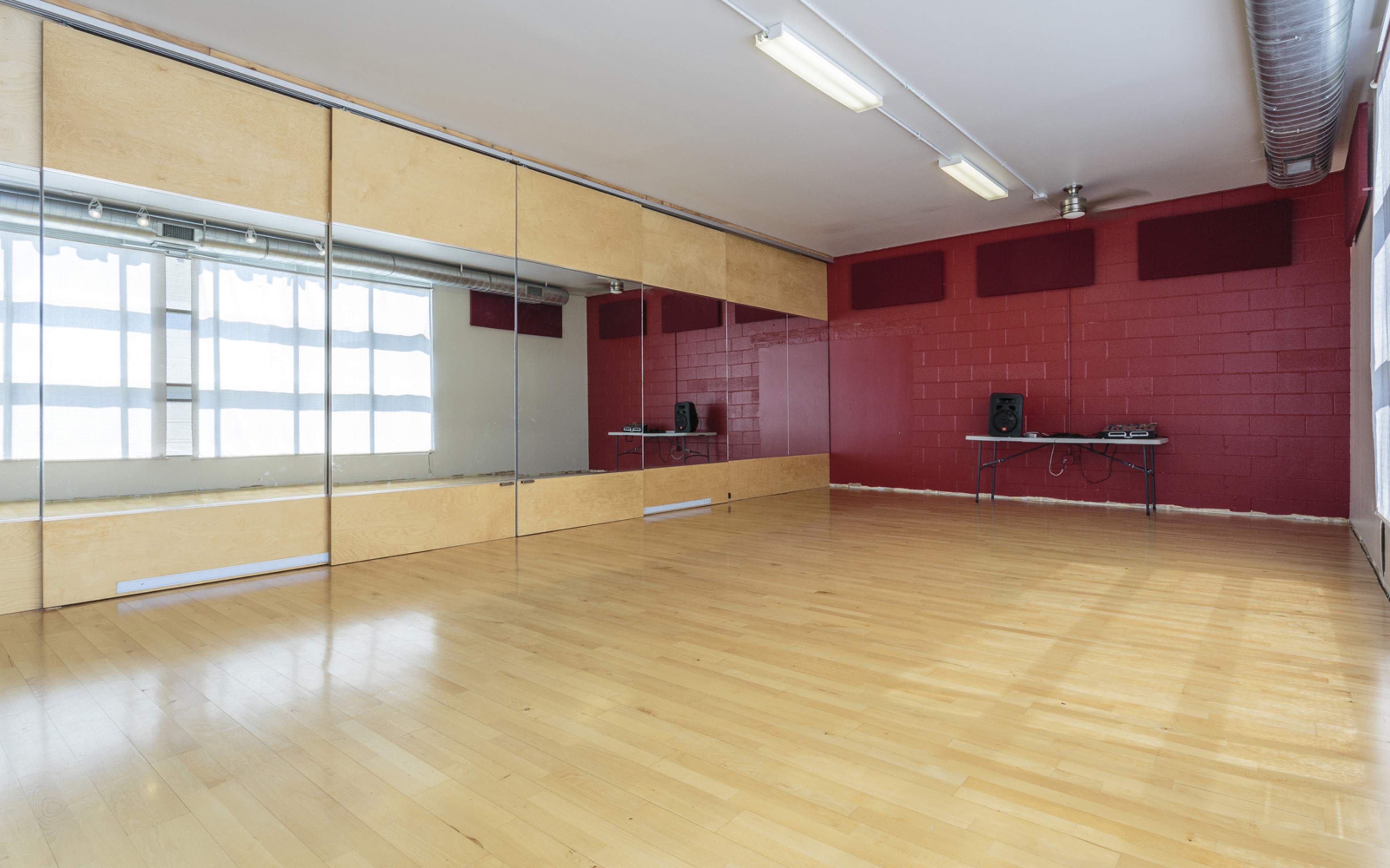 A dance studio featuring a wooden floor, large mirrors along one wall, and red-painted walls with a table and speaker setup in the corner.