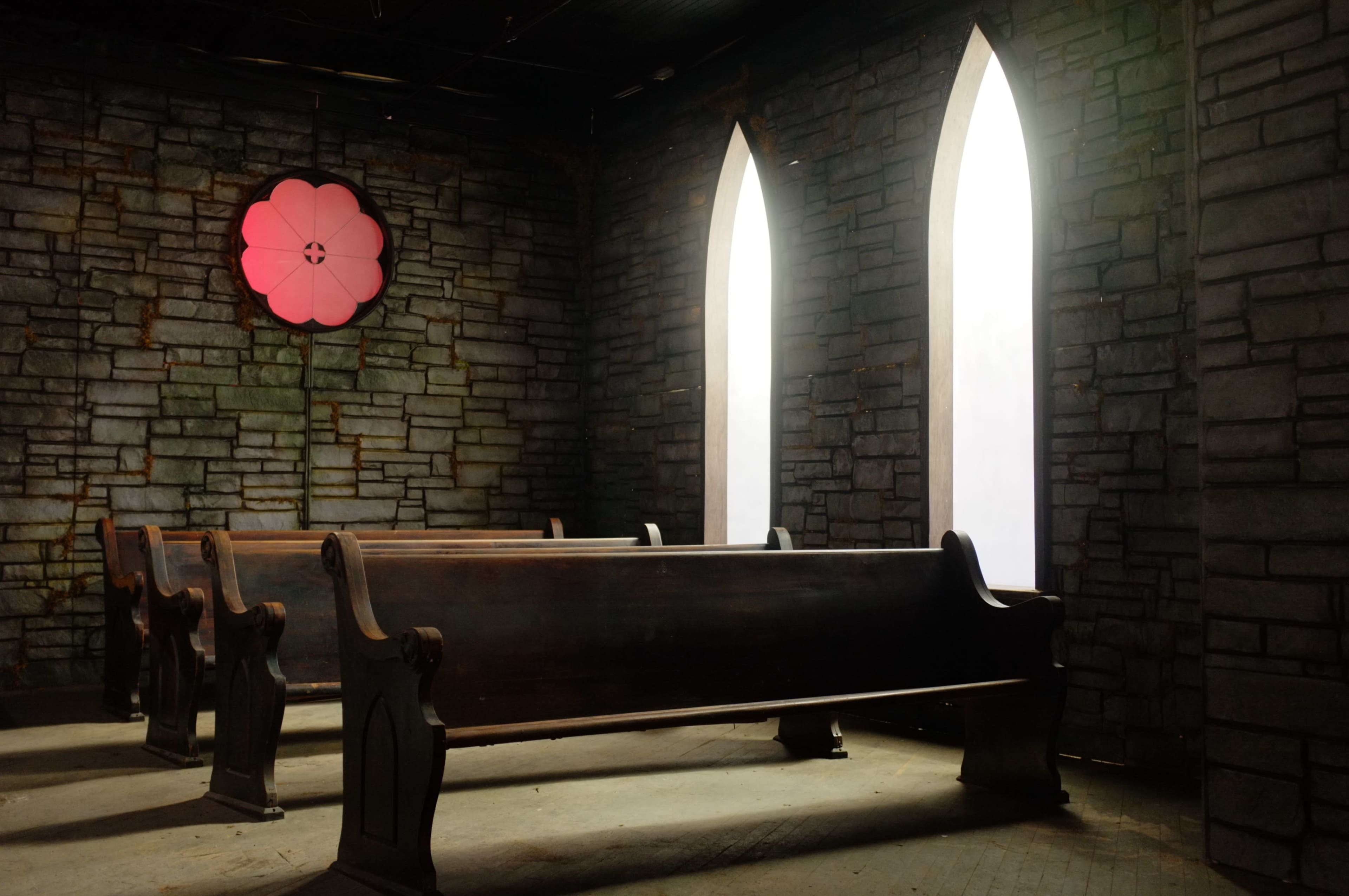 The image shows a dimly lit room with stone walls, featuring two tall arched windows and wooden pews arranged in front.