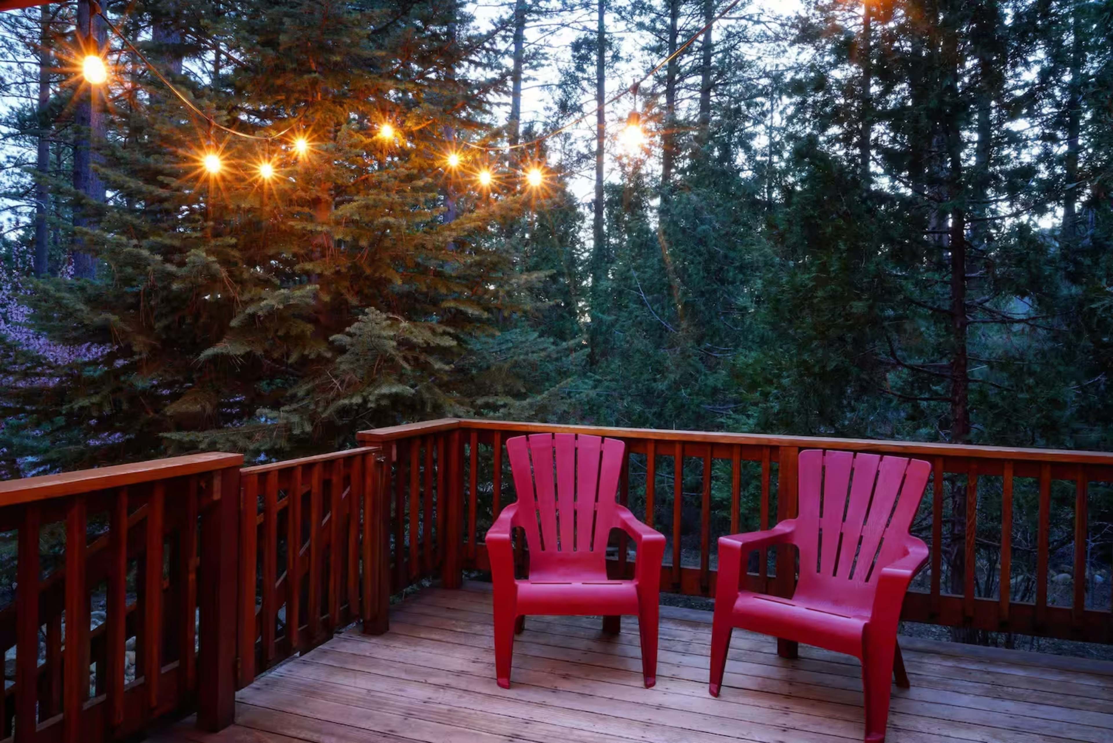 Two red Adirondack chairs are placed on a wooden deck surrounded by tall trees and illuminated by string lights at dusk.