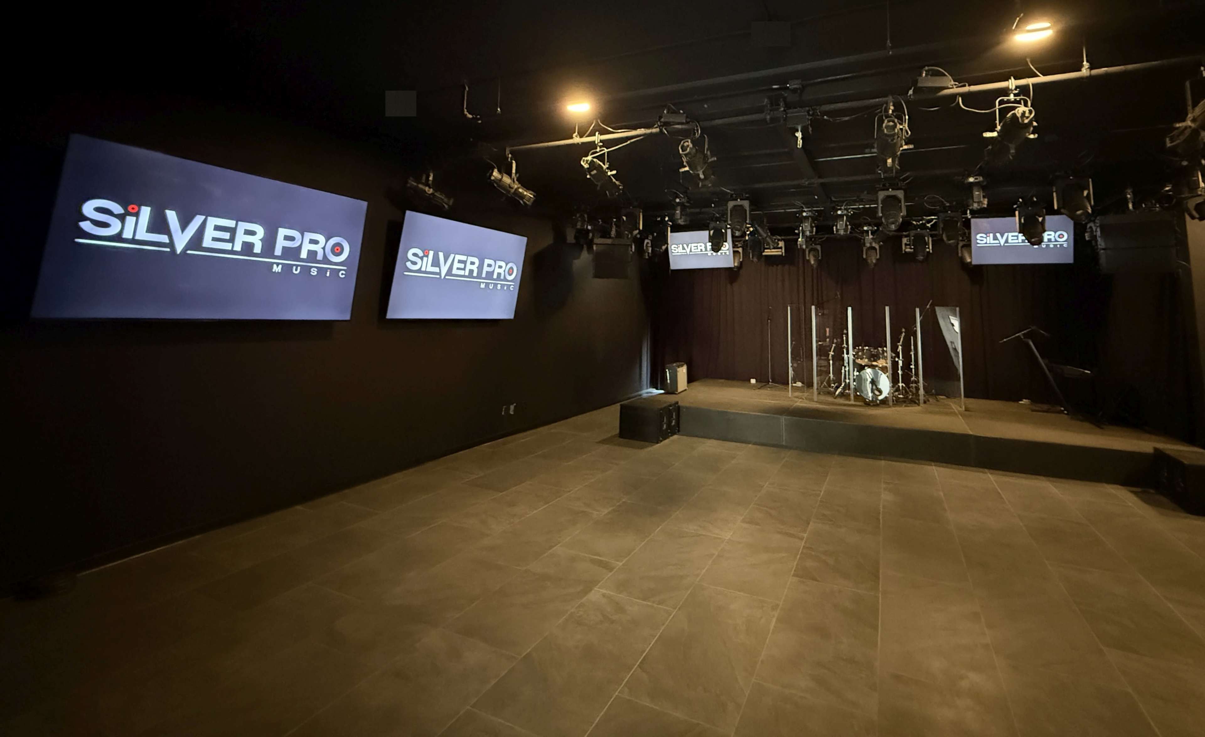 The image shows a performance venue with a dark interior, featuring a stage set up with a drum kit, hanging lights, and three screens displaying the logo "SILVER PRO MUSIC."