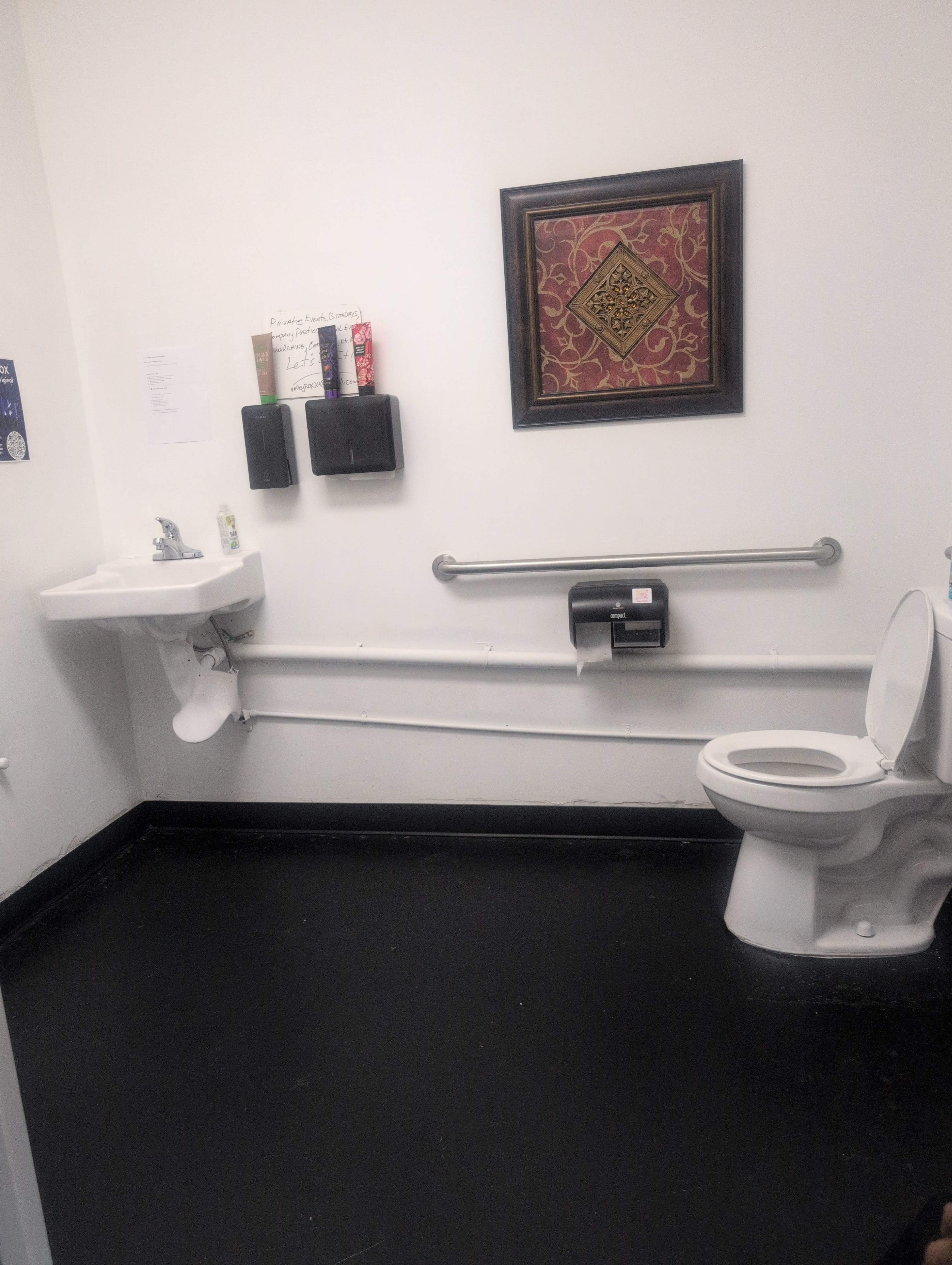 The image shows a small bathroom with a sink on the left, a toilet on the right, and wall-mounted dispensers for soap and paper towels.