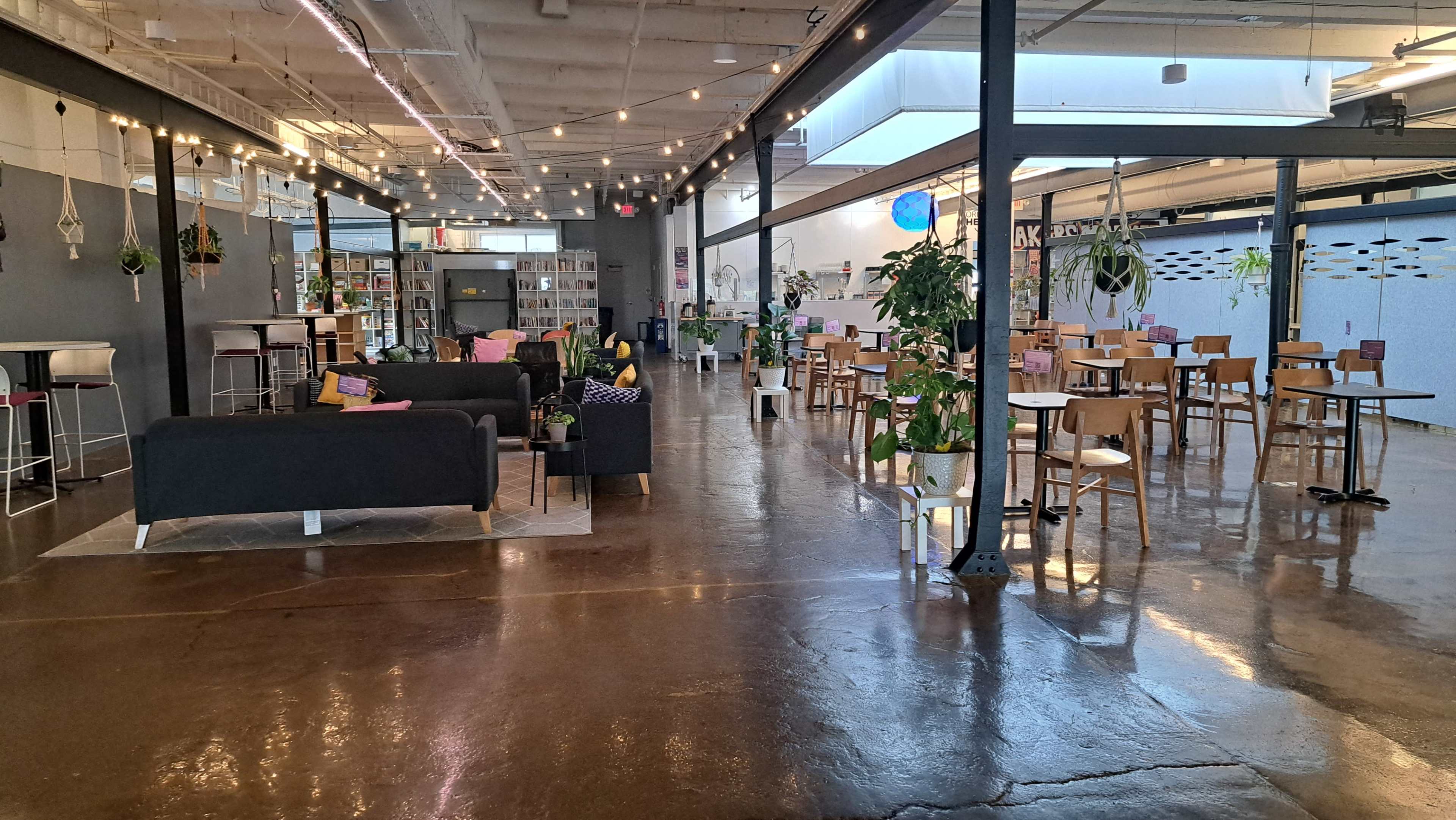 The image shows a spacious, modern co-working area featuring sofas, tables, and plants, with a library area in the background.
