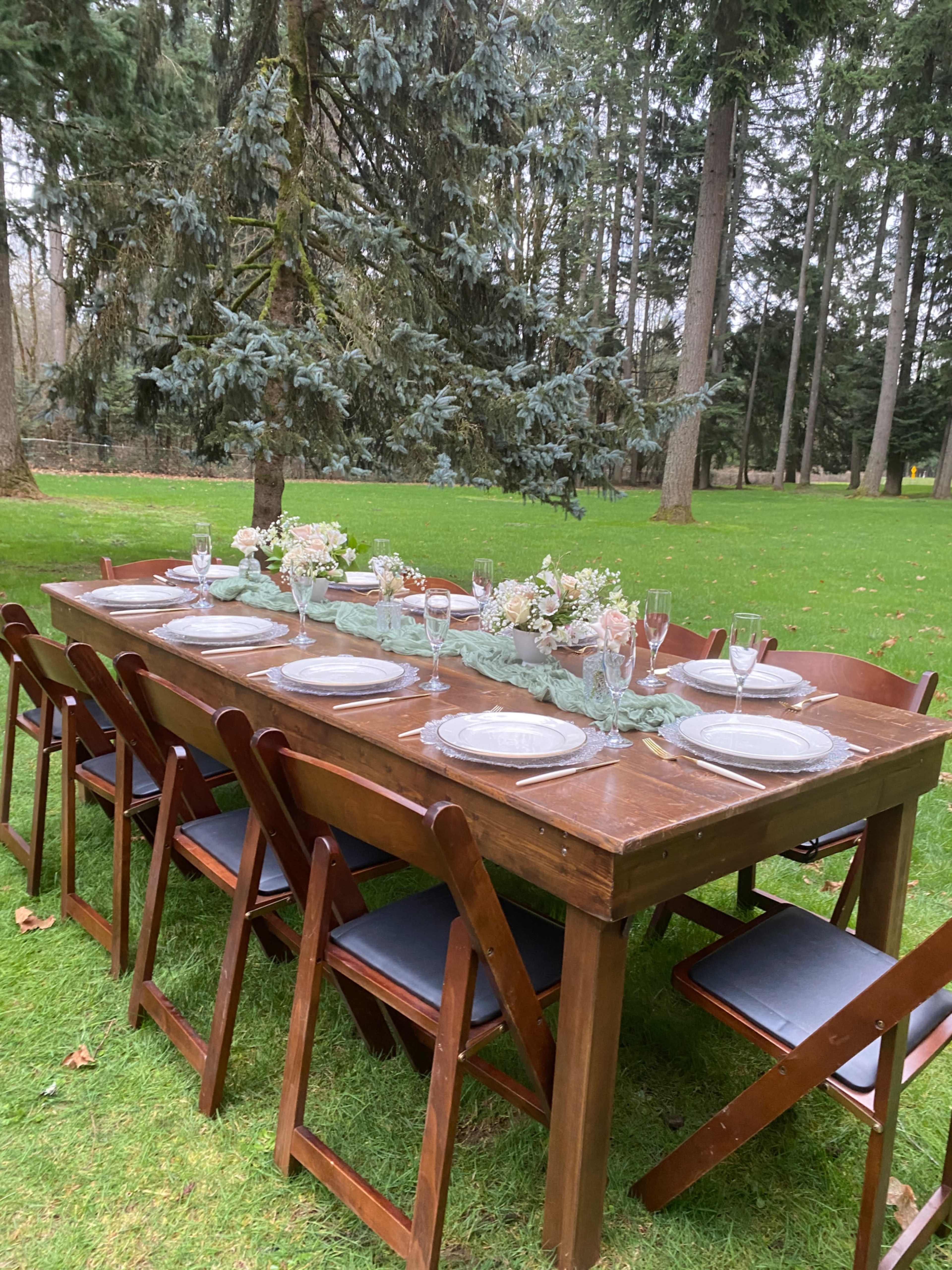 A long wooden dining table is set outdoors on a grassy area, decorated with flowers and surrounded by wooden chairs.