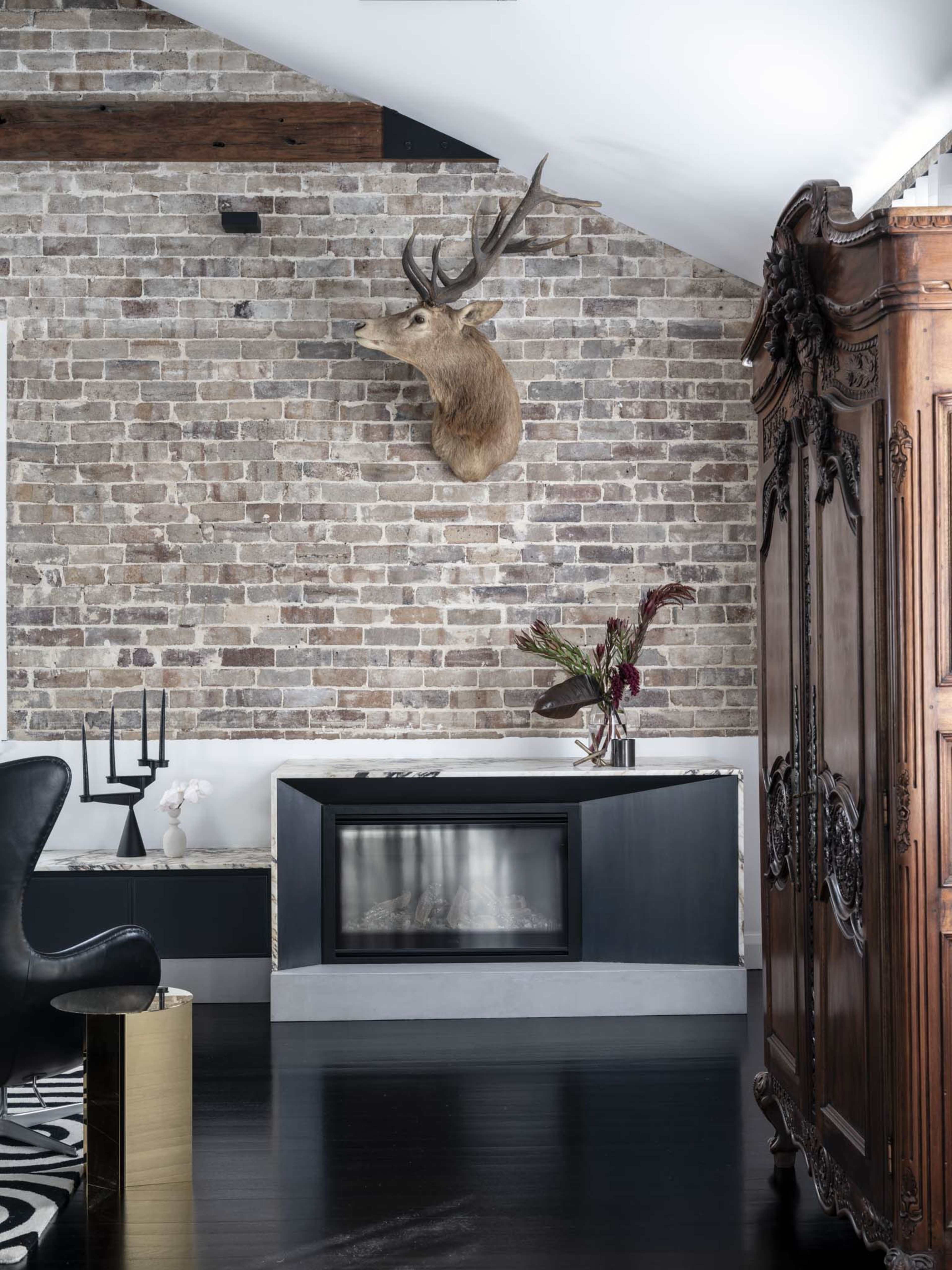The image shows a modern living space featuring a brick wall, a mounted deer head, a minimalist black fireplace, and an ornate wooden cabinet.