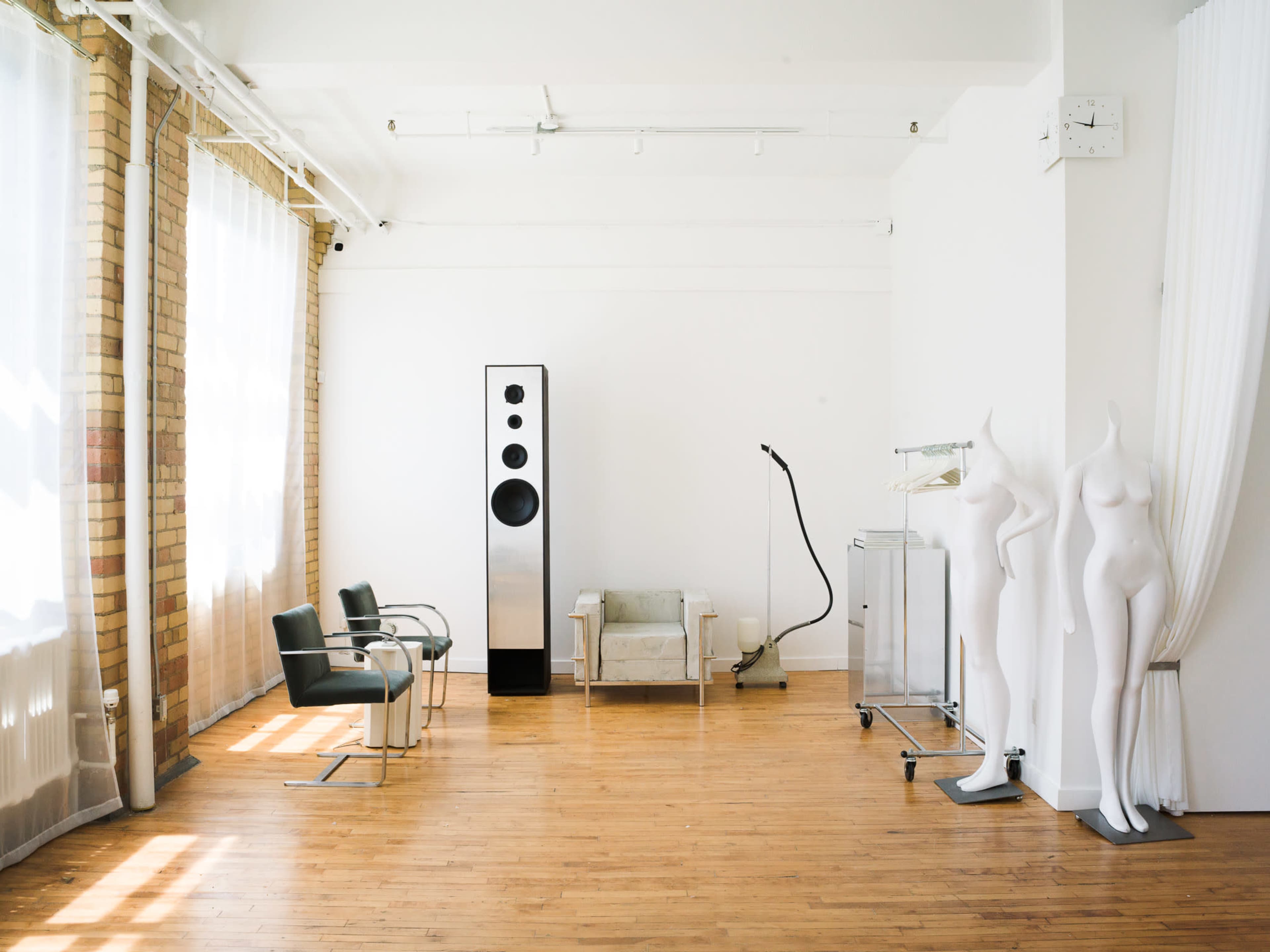 The image shows a bright, minimalist interior space featuring two chairs, a modern speaker, a mannequin, and a medical cart against white walls and wooden flooring.