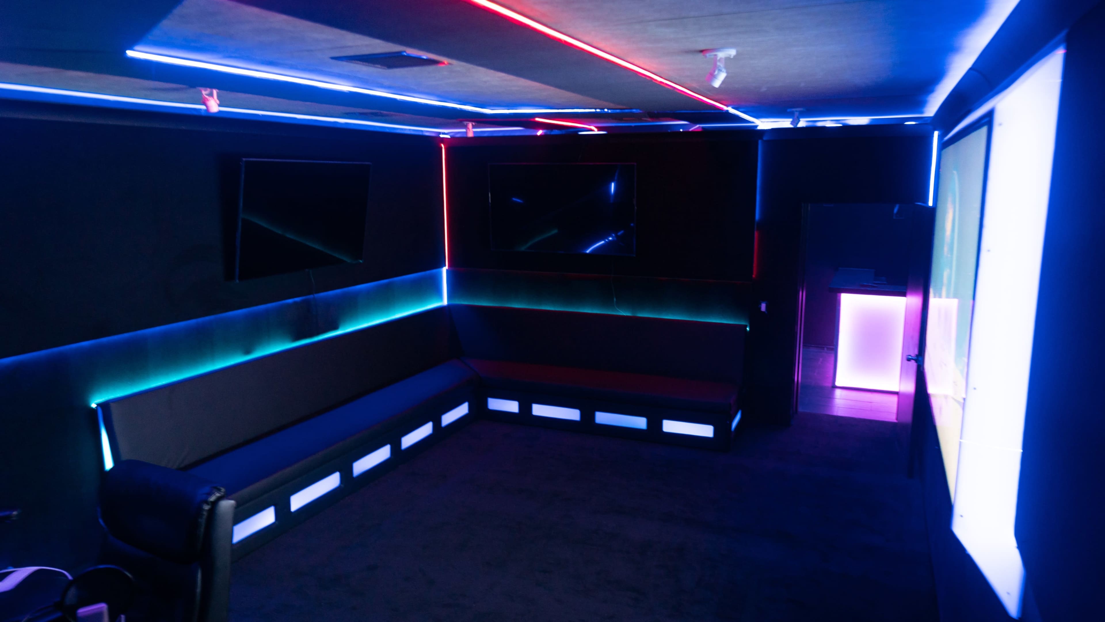 A dimly lit room with black walls and illuminated LED strips in blue and red, along with two large screens mounted on the walls.
