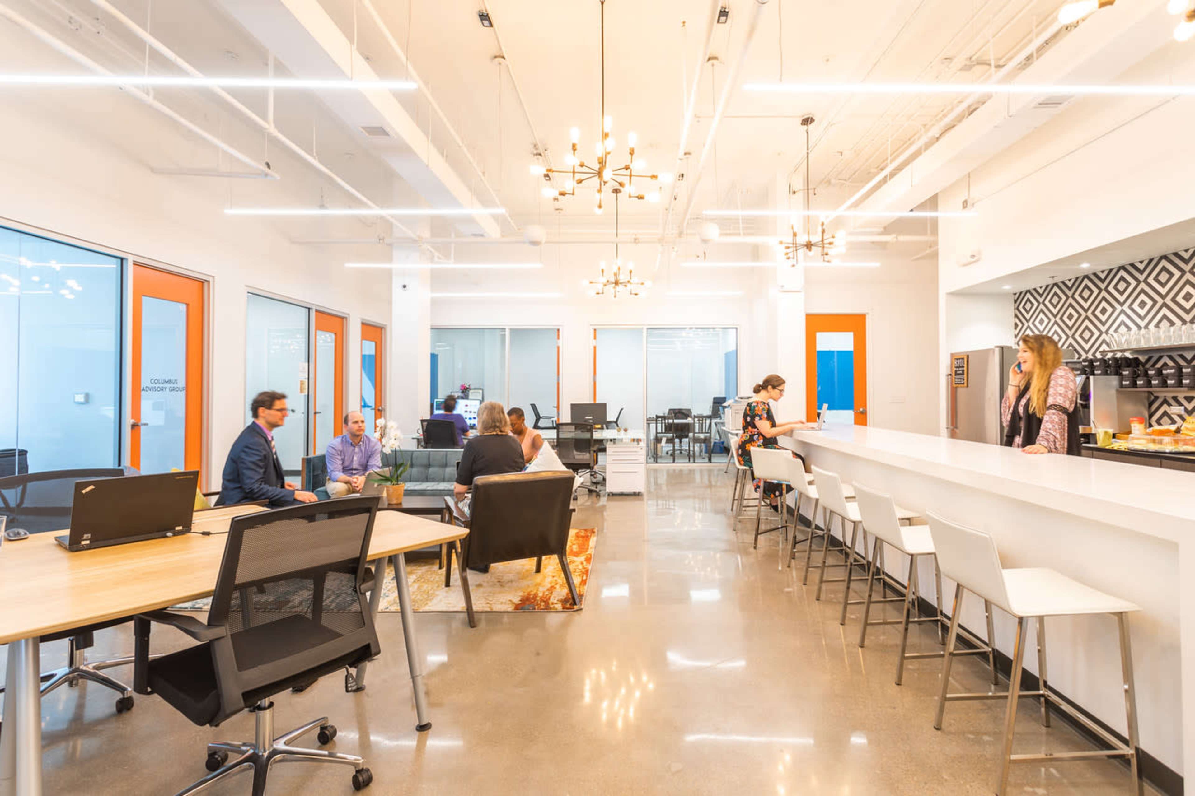 The image shows a modern office space with people engaged in discussions, a central bar area, and workstations visible in the background.