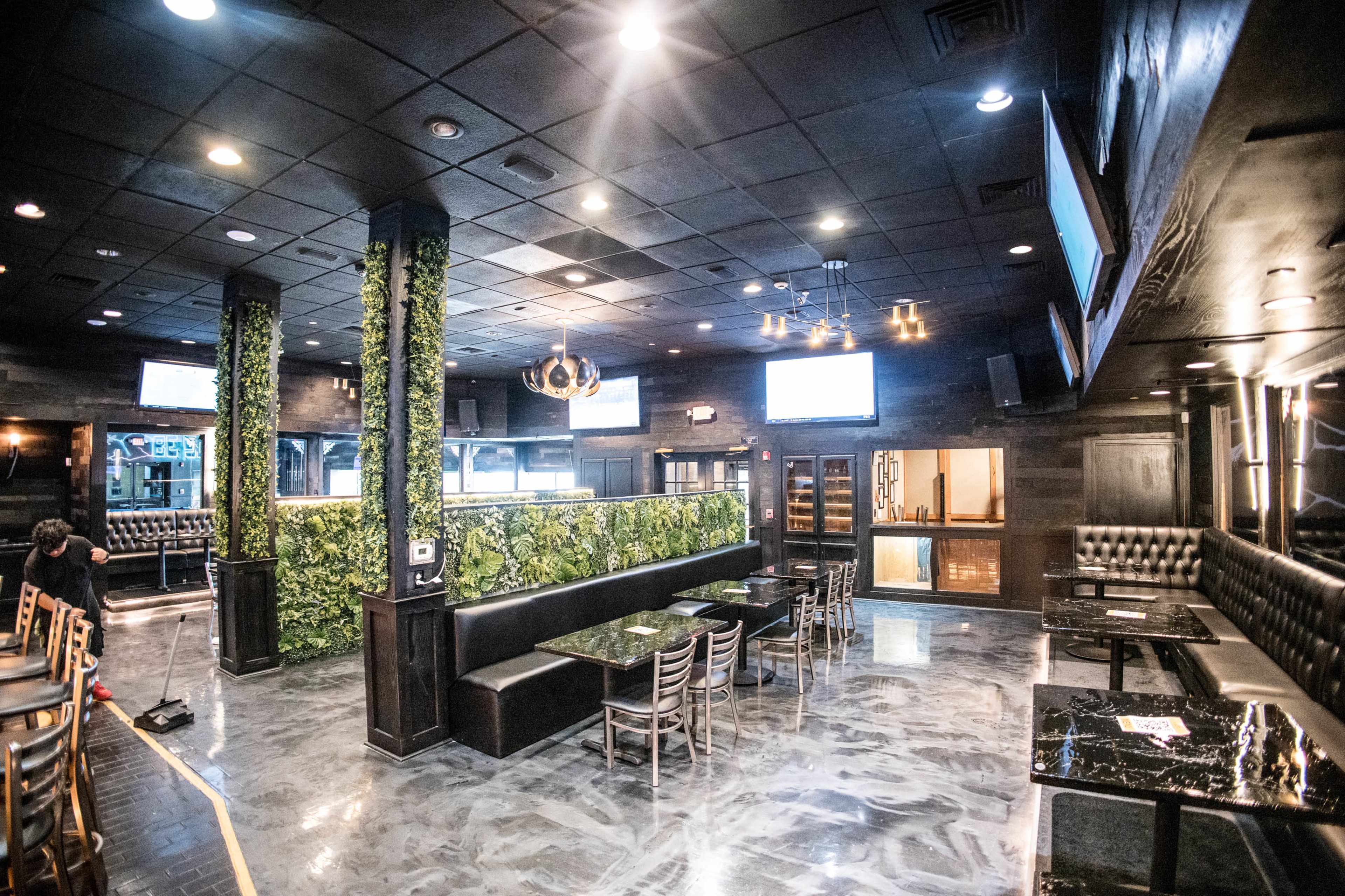The image shows a modern bar interior with dark furnishings, green plant walls, and multiple television screens mounted on the walls.