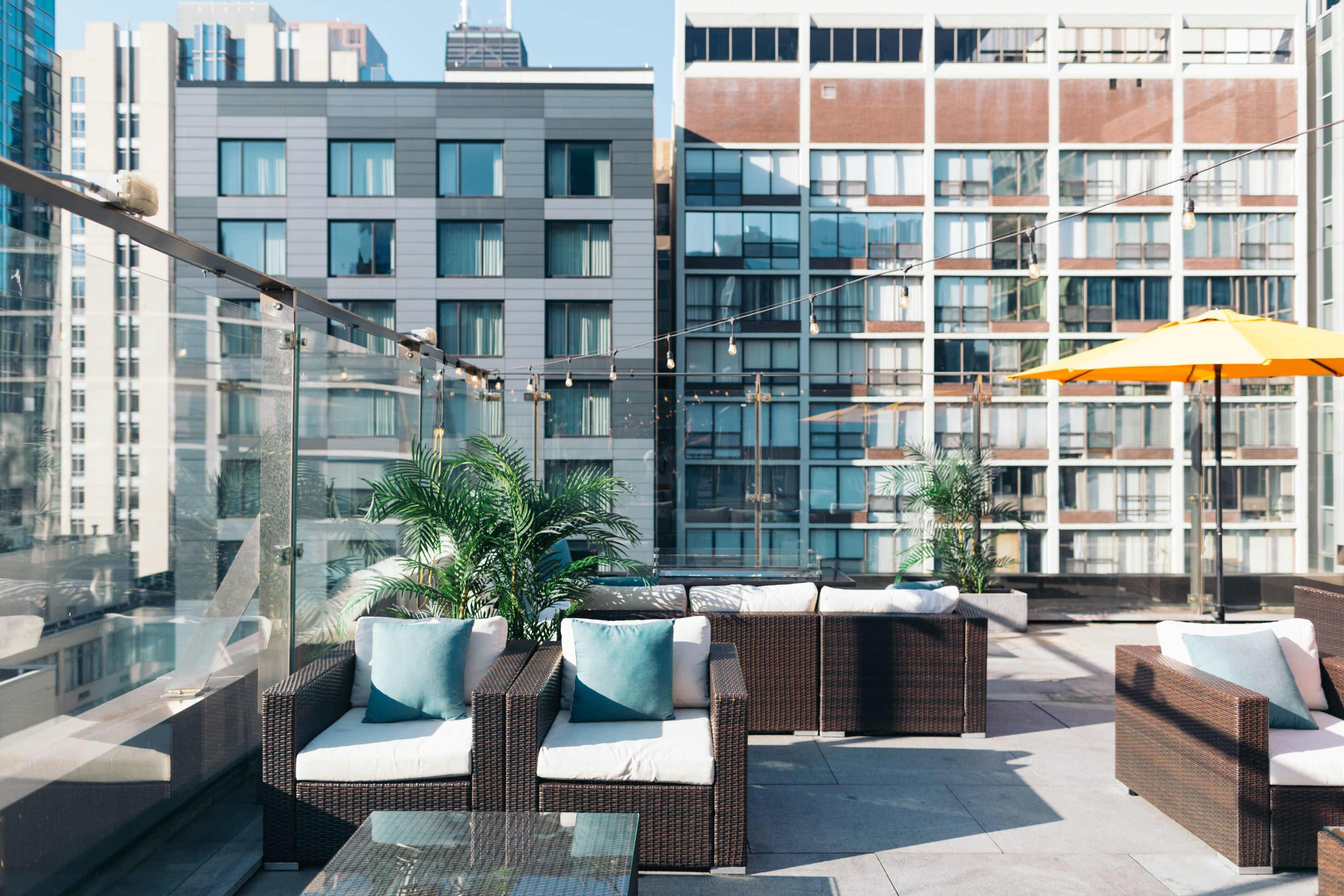 The rooftop area features modern wicker furniture with blue cushions, surrounded by tall buildings and a bright orange umbrella.