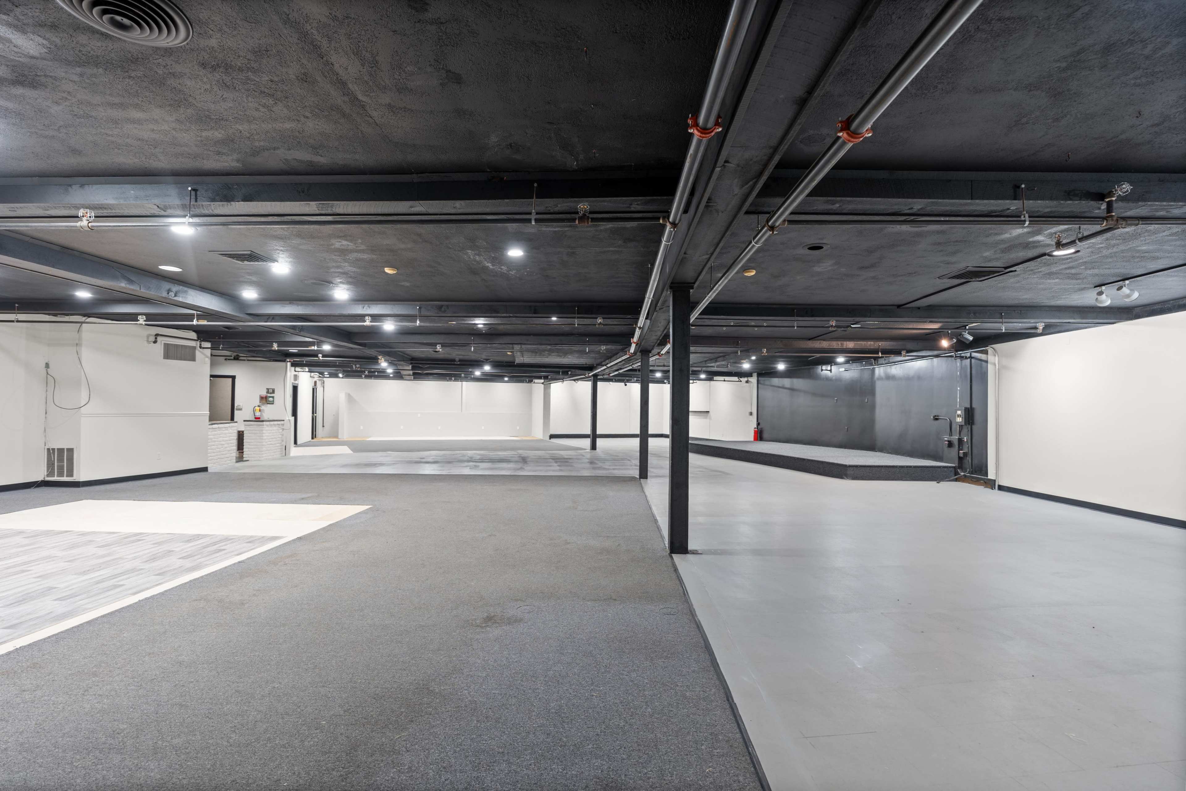 The image shows a large, empty commercial space with dark ceilings, concrete and carpeted floors, and multiple columns.