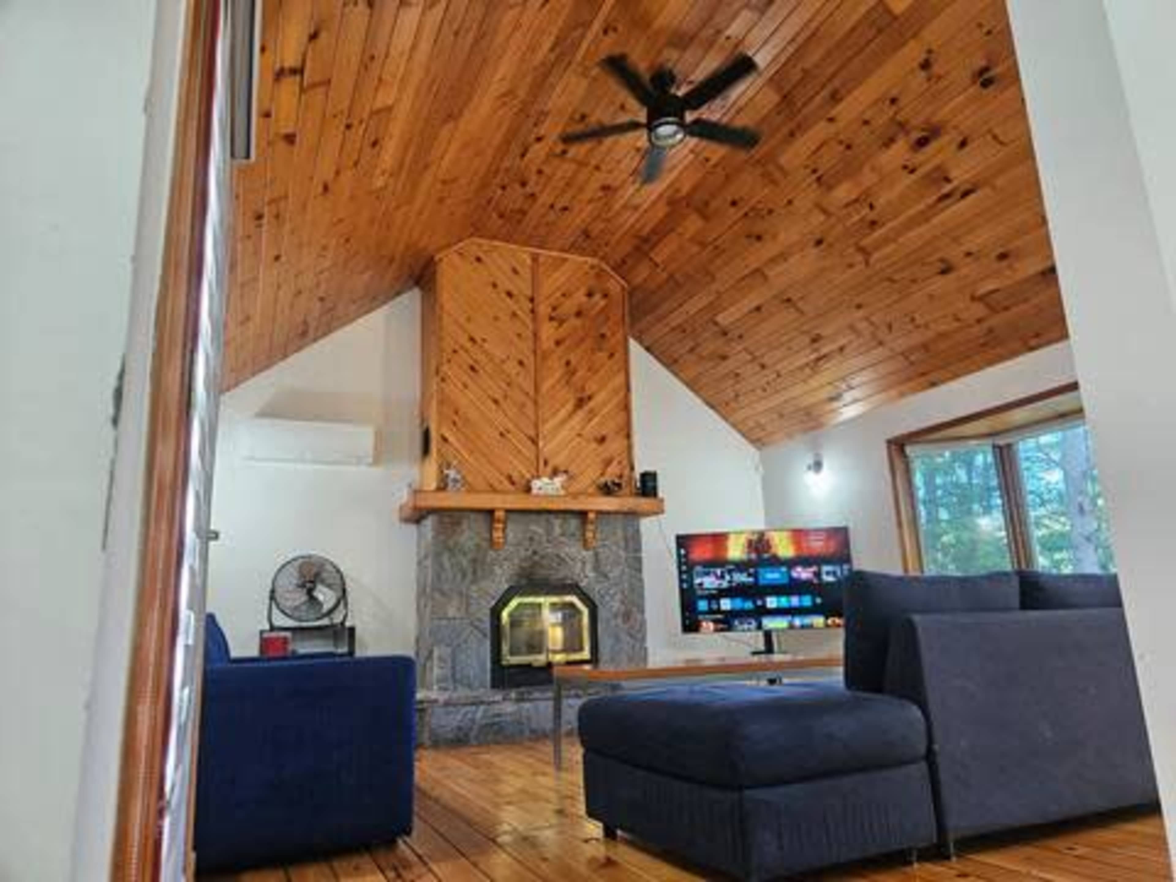 The image shows a cozy living room with a wooden ceiling, a stone fireplace, a large television mounted on the wall, and two dark sofas arranged around a coffee table.