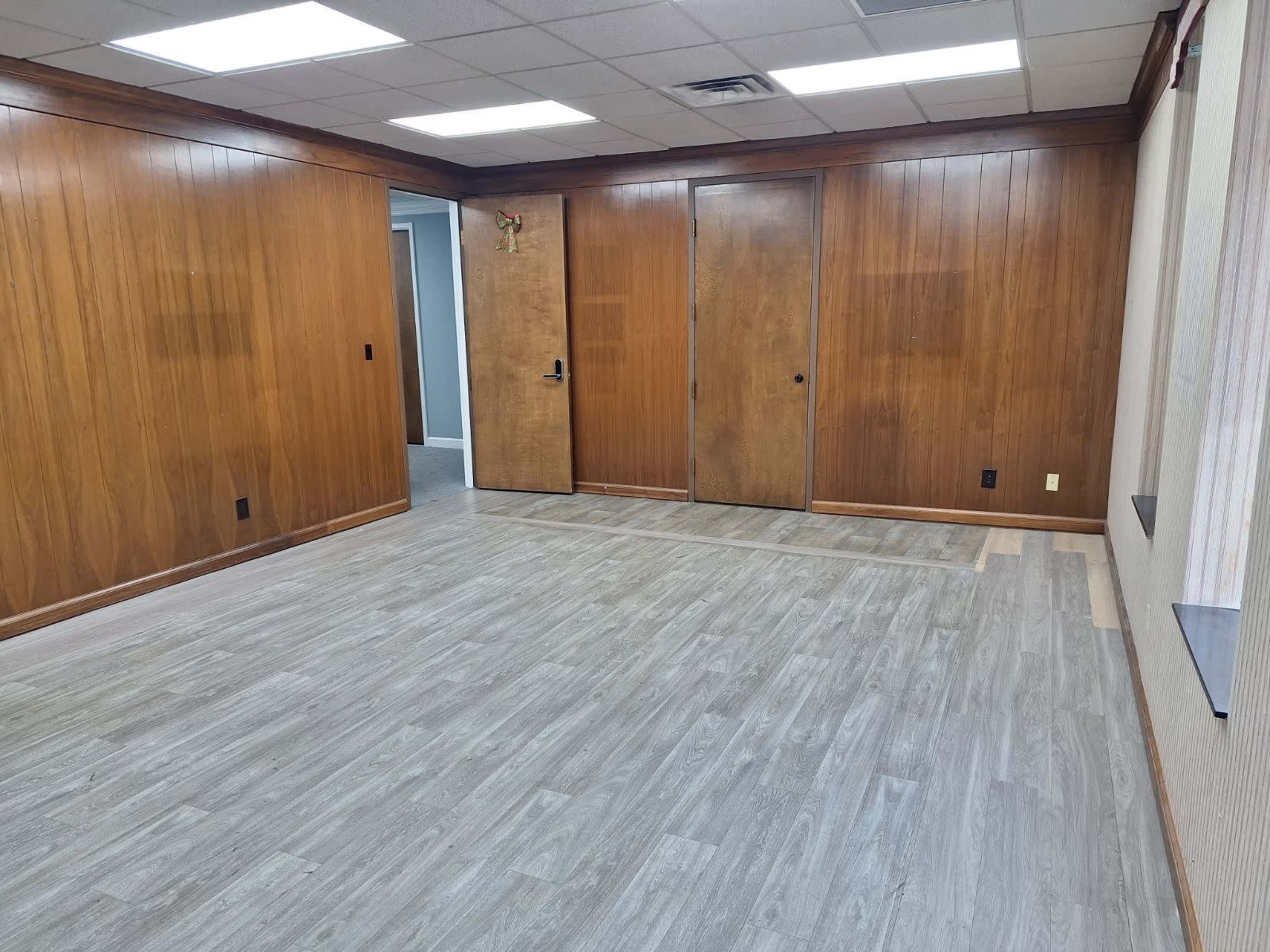 The image shows an empty room with wooden paneling on the walls and a light-colored laminate floor.