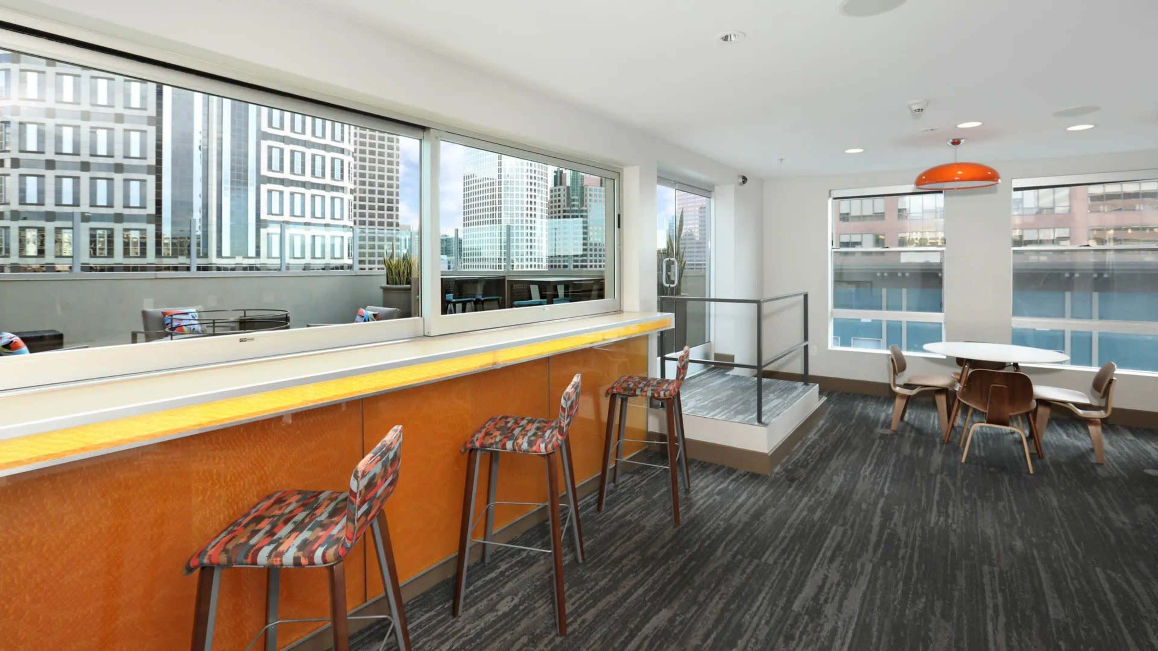 A modern interior space with a bar area featuring high stools, large windows overlooking city buildings, and a round dining table.