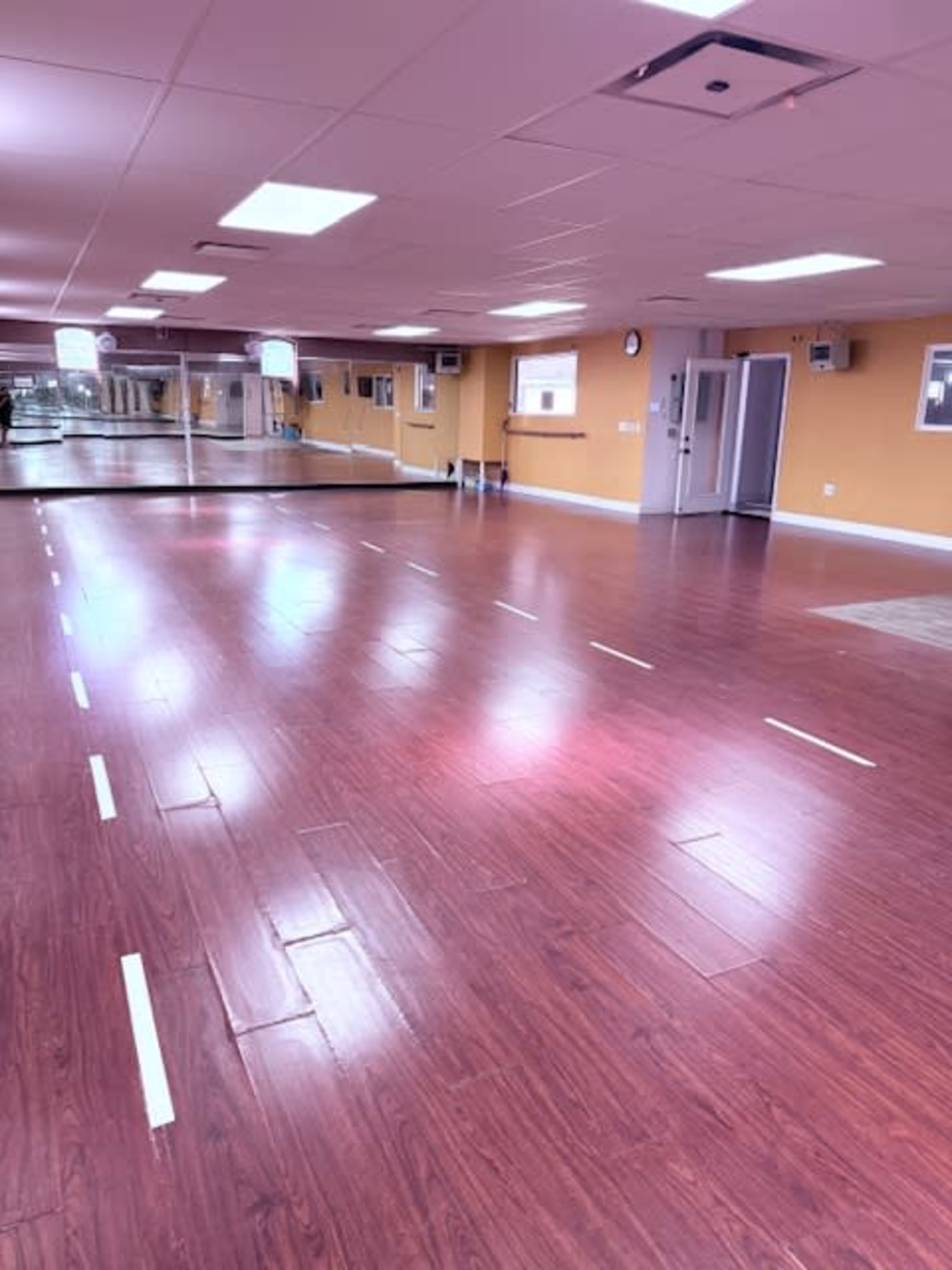 The image shows a spacious, empty studio with wooden flooring, marked lines on the floor, and mirrors along one wall.