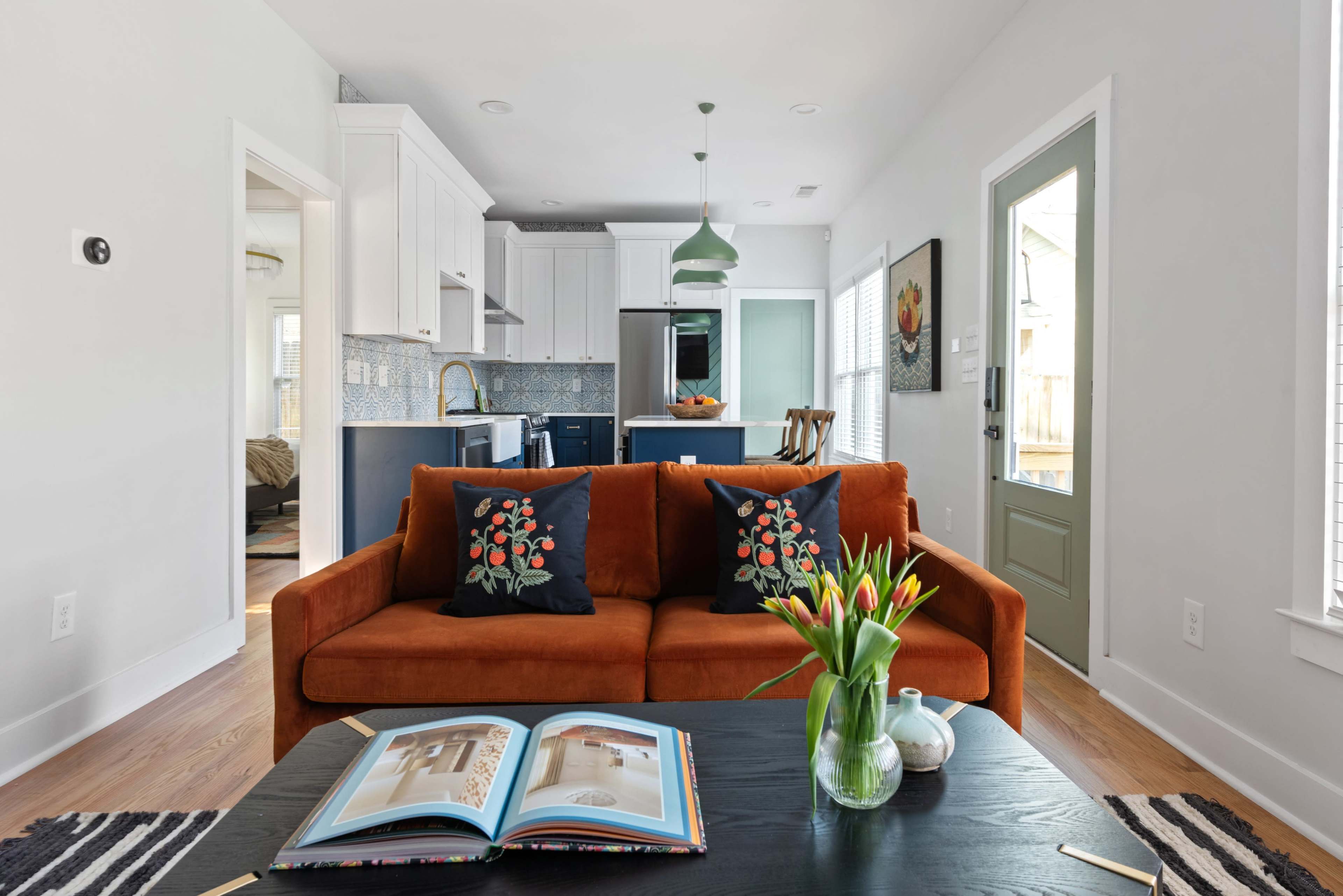A bright living room with an orange couch adorned with floral pillows, a black coffee table, and a kitchen area visible in the background.