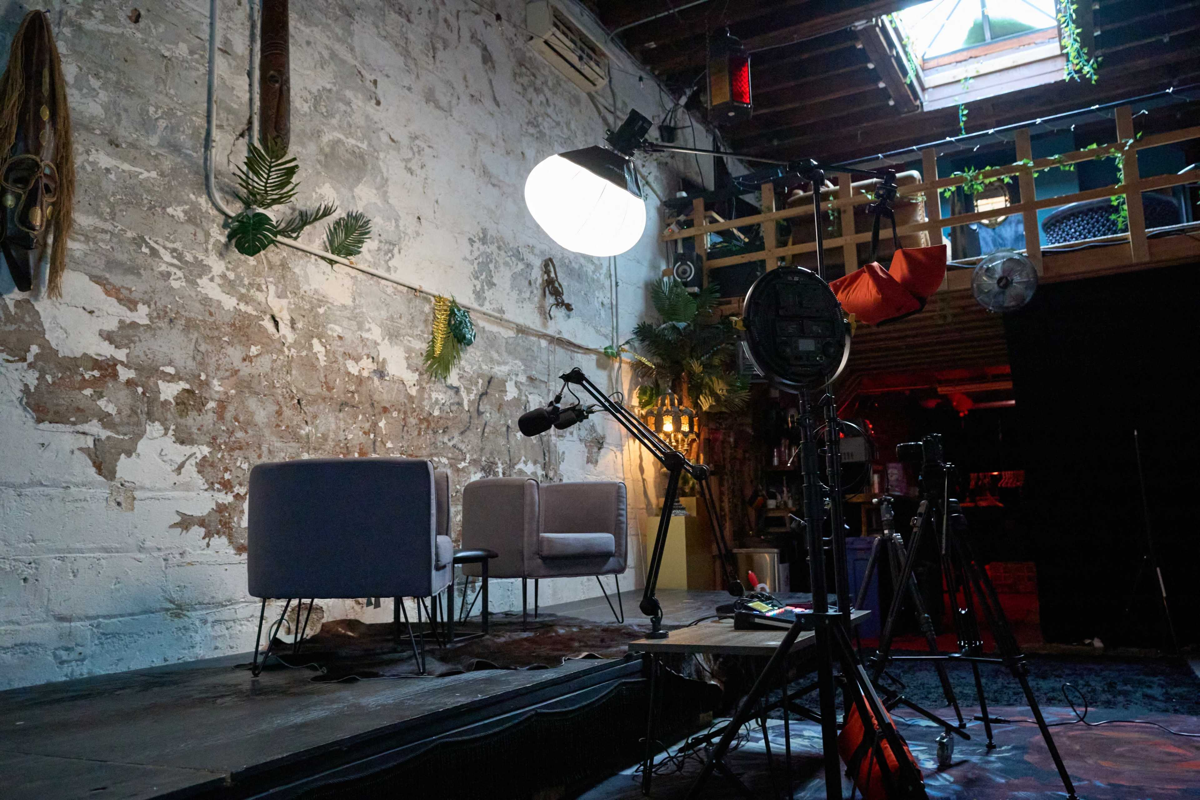 The image shows a rustic interior of a studio space with two chairs on a wooden platform, surrounded by recording equipment and decorative plants.