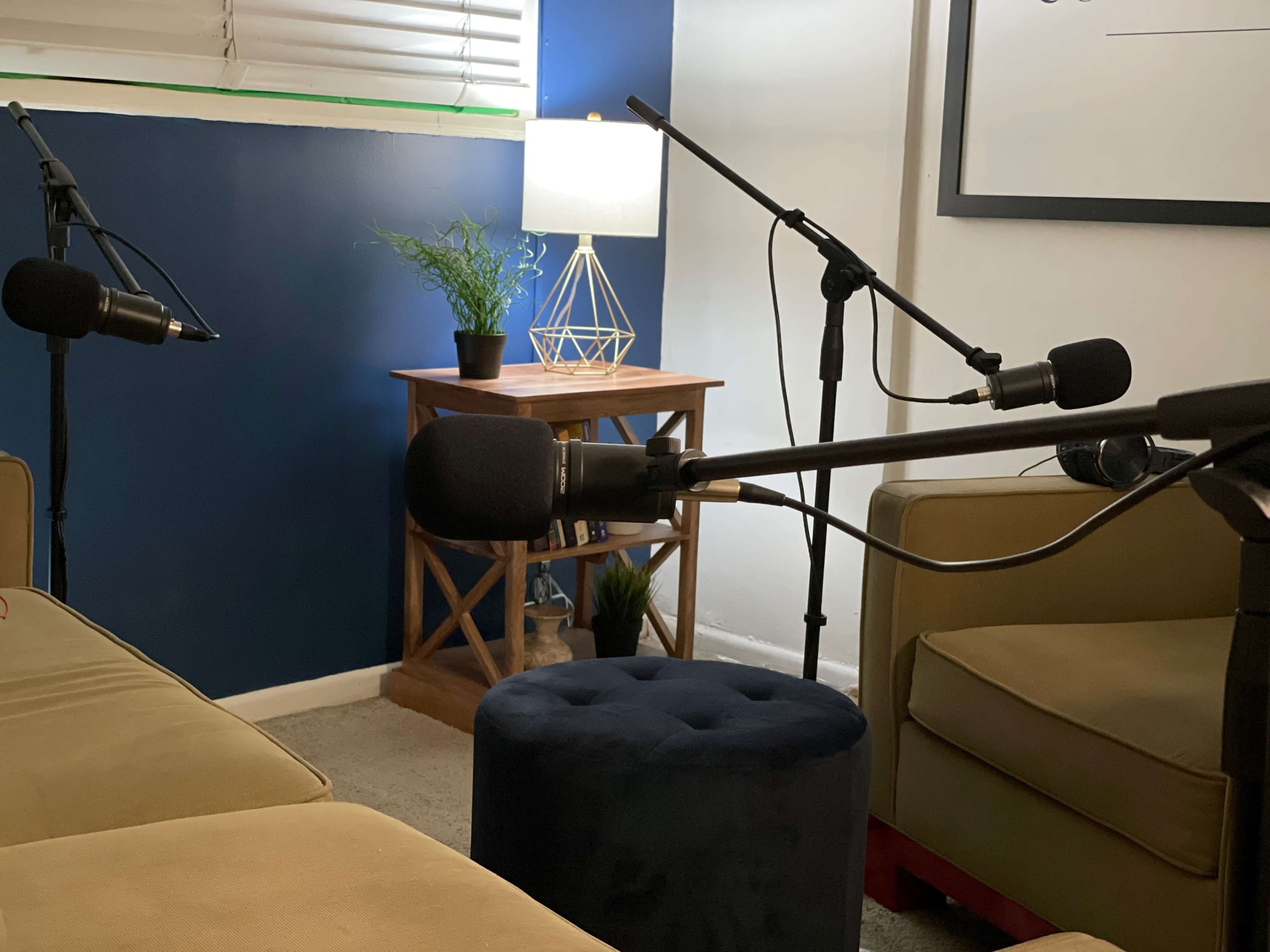 A small recording space with microphones set up around a pair of green couches and a blue ottoman, accompanied by a lamp and a potted plant on a wooden table.