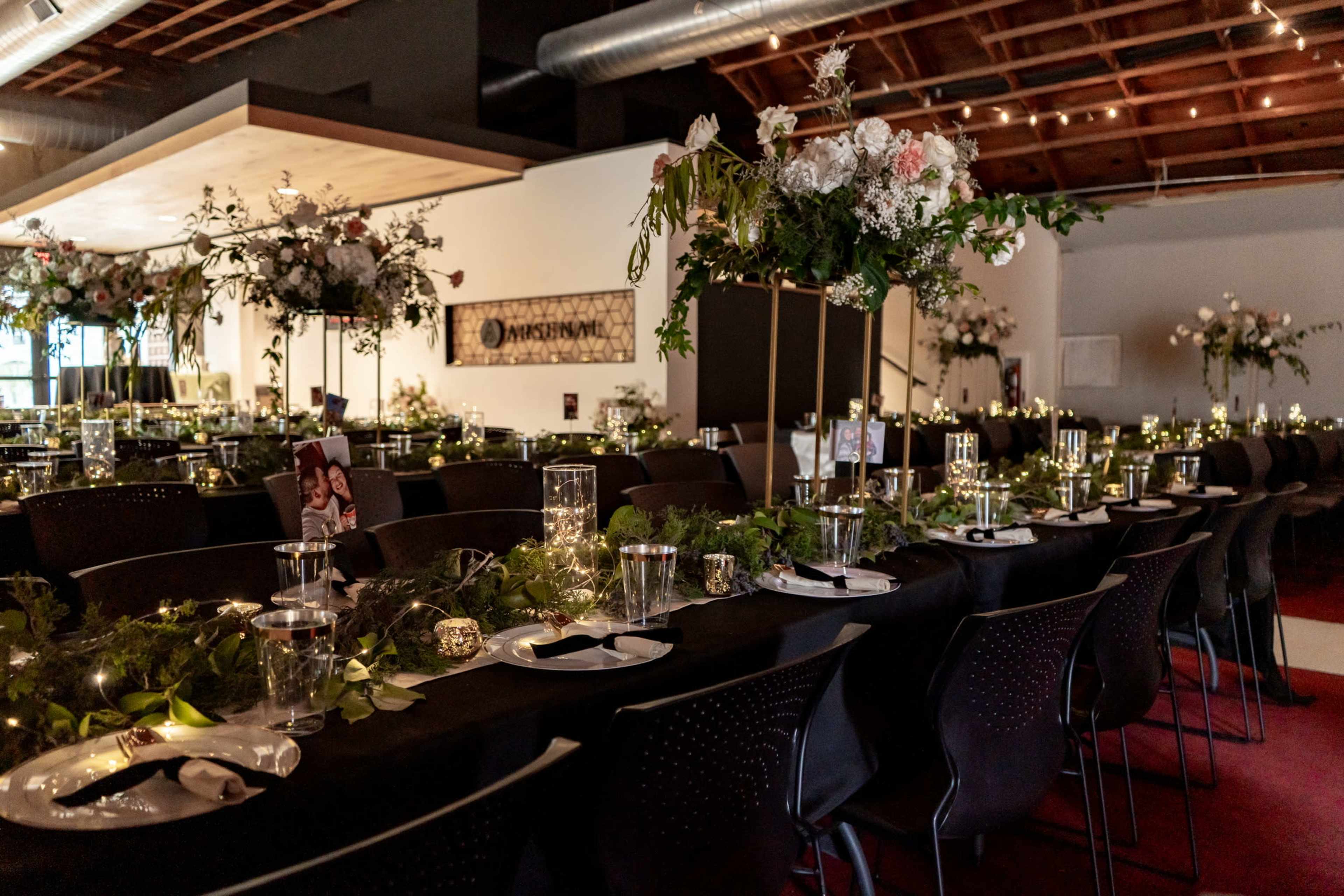 A beautifully arranged dining setup features long tables adorned with greenery, candles, and floral centerpieces in a well-lit event space.