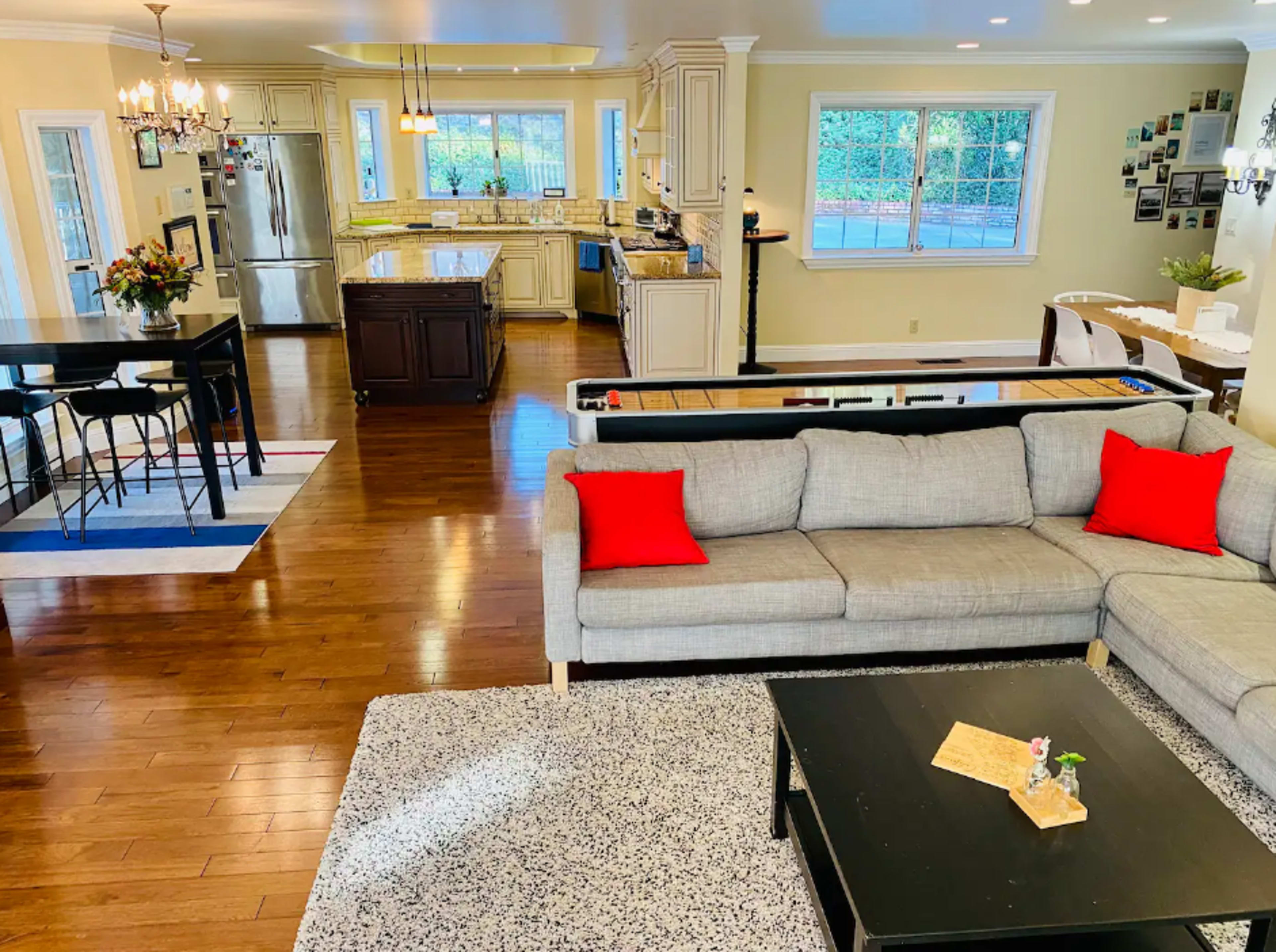 The image shows a spacious, modern living room with a gray sectional sofa, a black coffee table, and an open kitchen in the background featuring light-colored cabinets and a large window.
