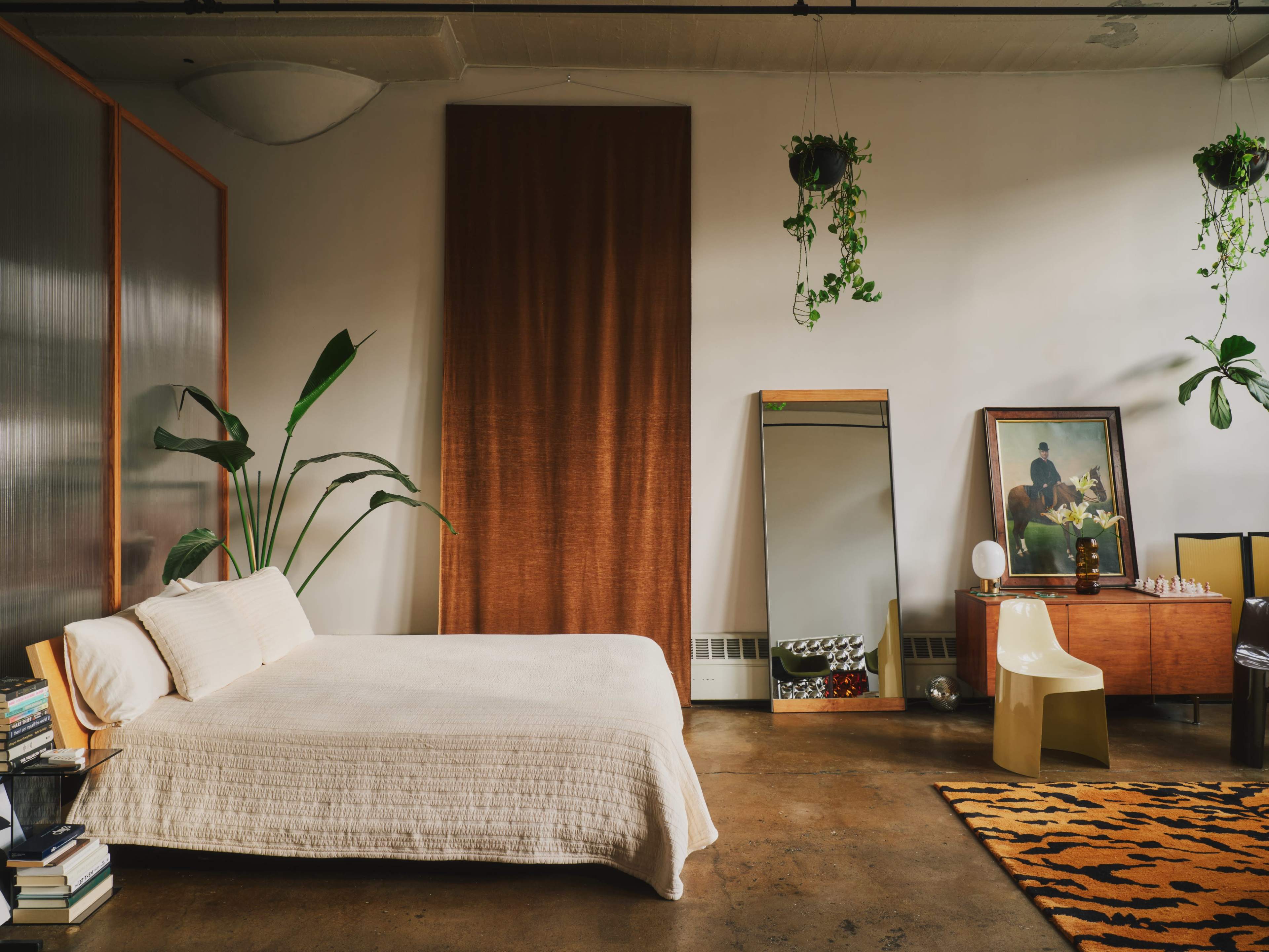 The image features a minimalist bedroom with a bed, large mirror, artwork, a wooden cabinet, and potted plants, set against a backdrop of concrete flooring and warm earthy tones.