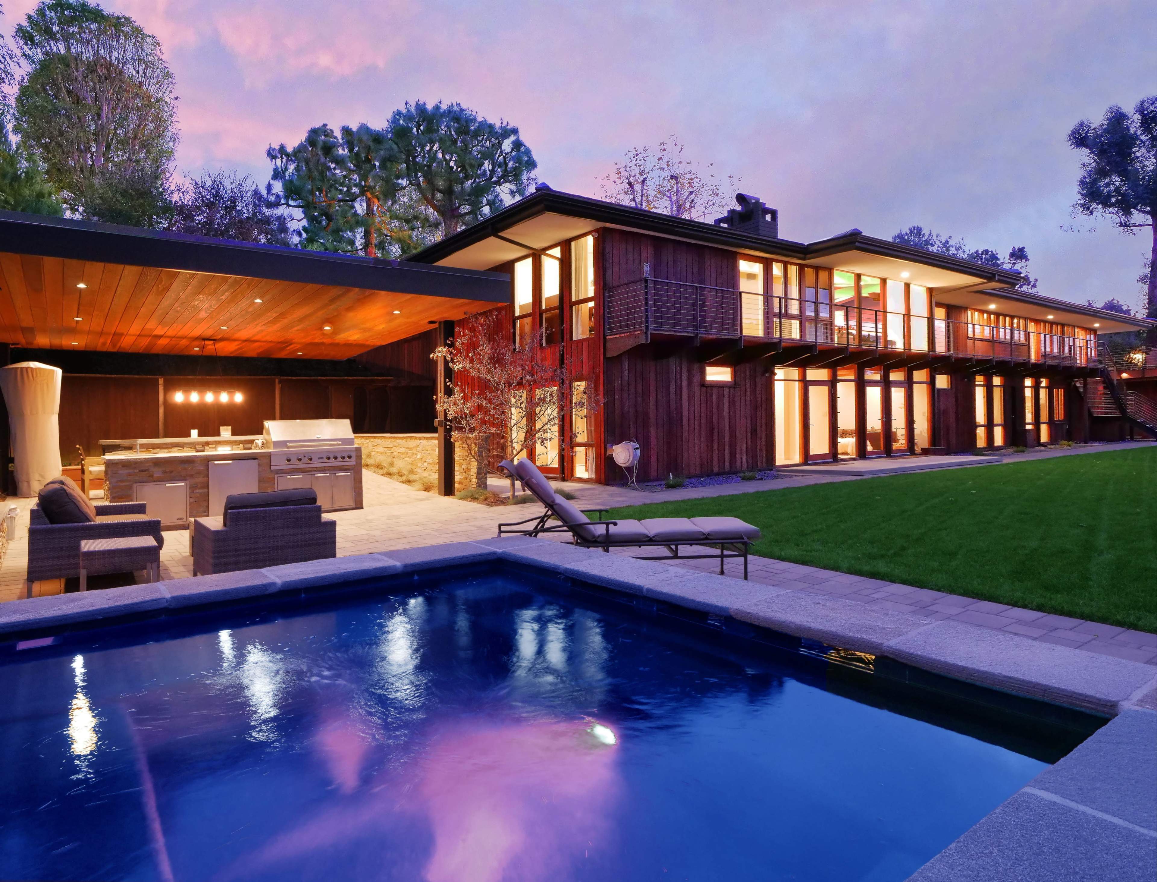 A modern house with large glass windows and a patio featuring an outdoor kitchen is set beside a swimming pool at dusk.