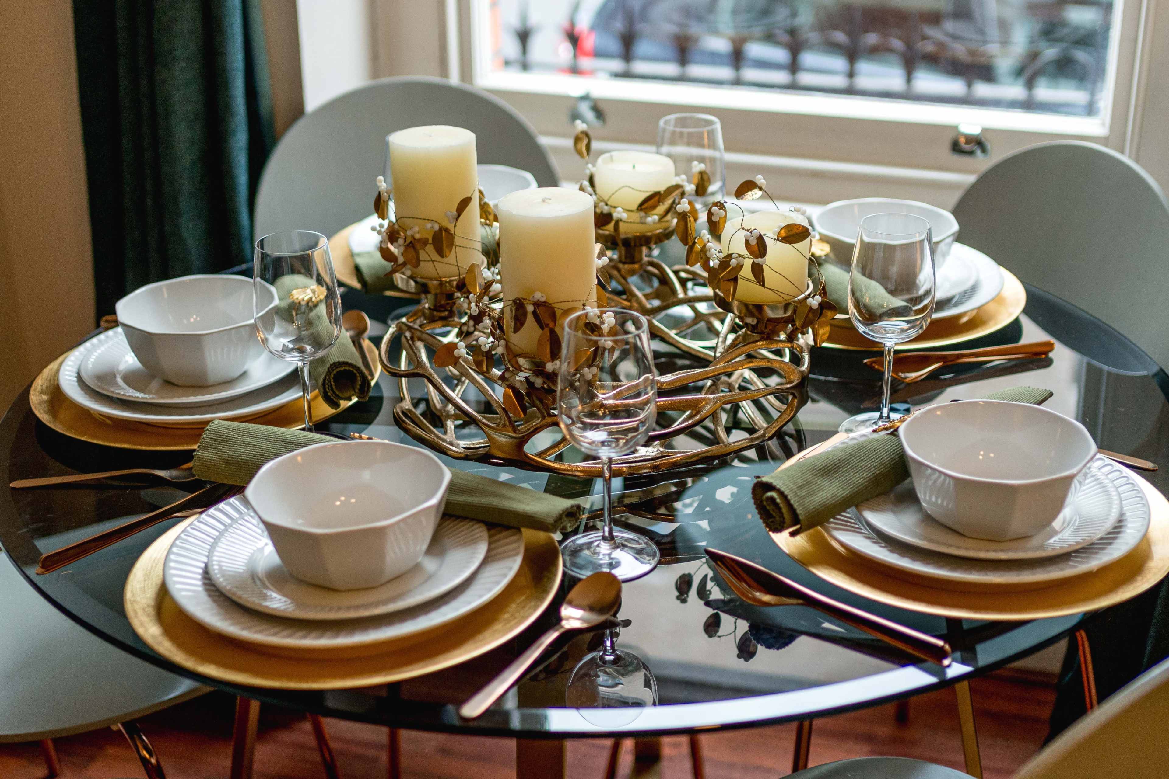 A round dining table is set with white bowls, glassware, gold-rimmed plates, decorative candles, and green napkins.