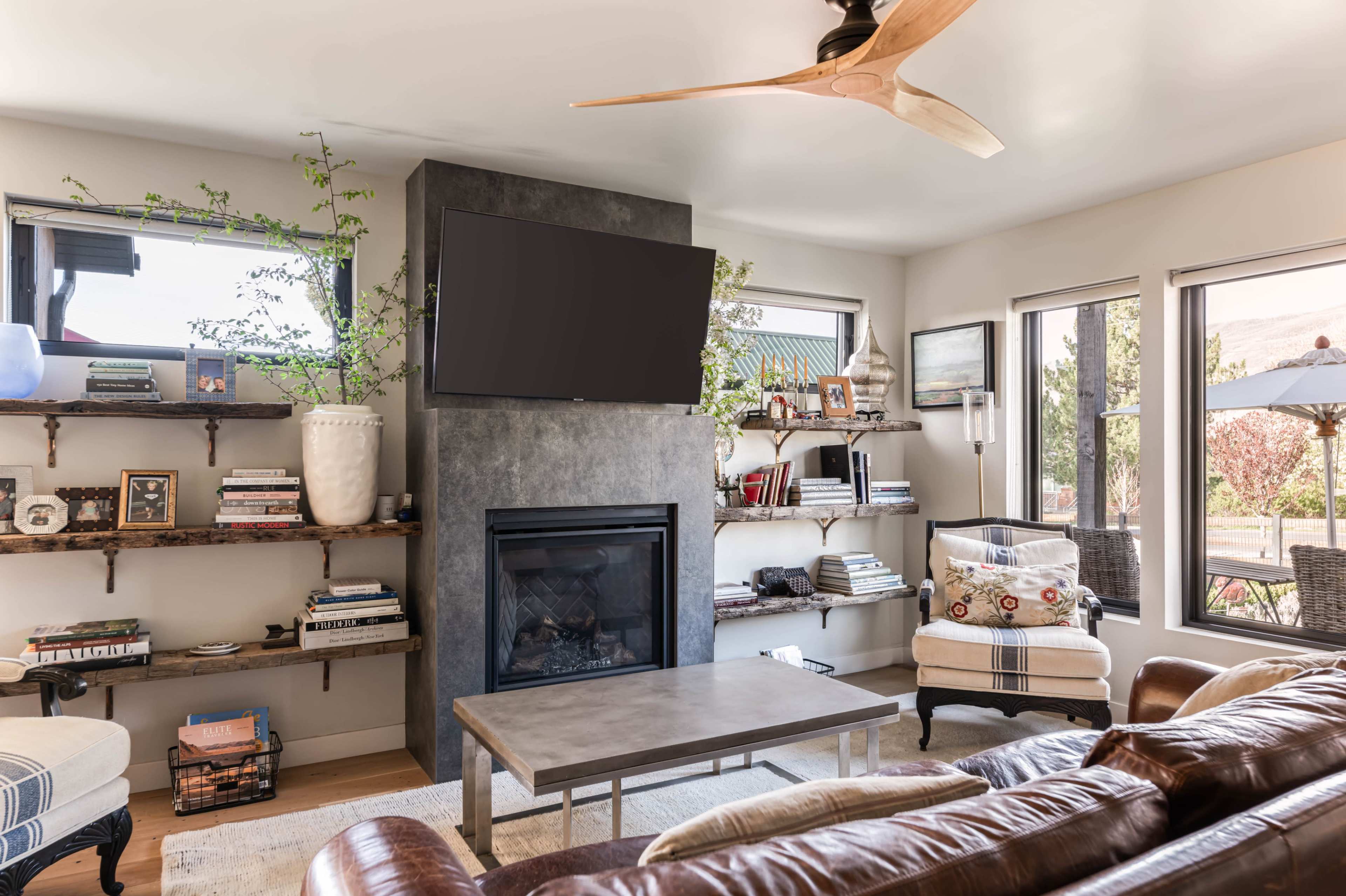 The image shows a modern living room featuring a sleek gray fireplace, a mounted television, shelves with books and decor, and comfortable seating arrangements near large windows.