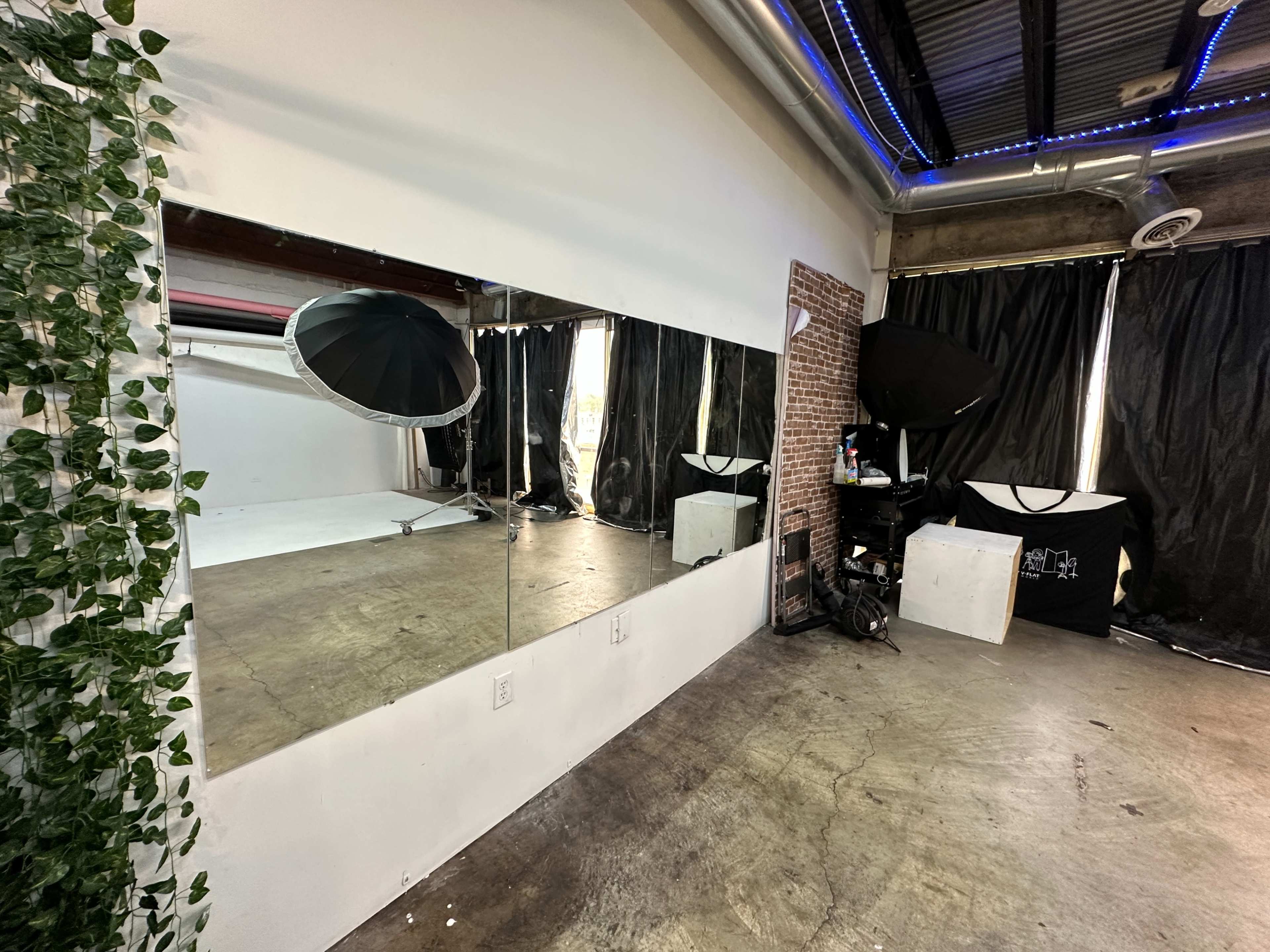 A spacious photography studio features a large mirror, a black umbrella softbox, and a backdrop area, with brick and black drapery accents.