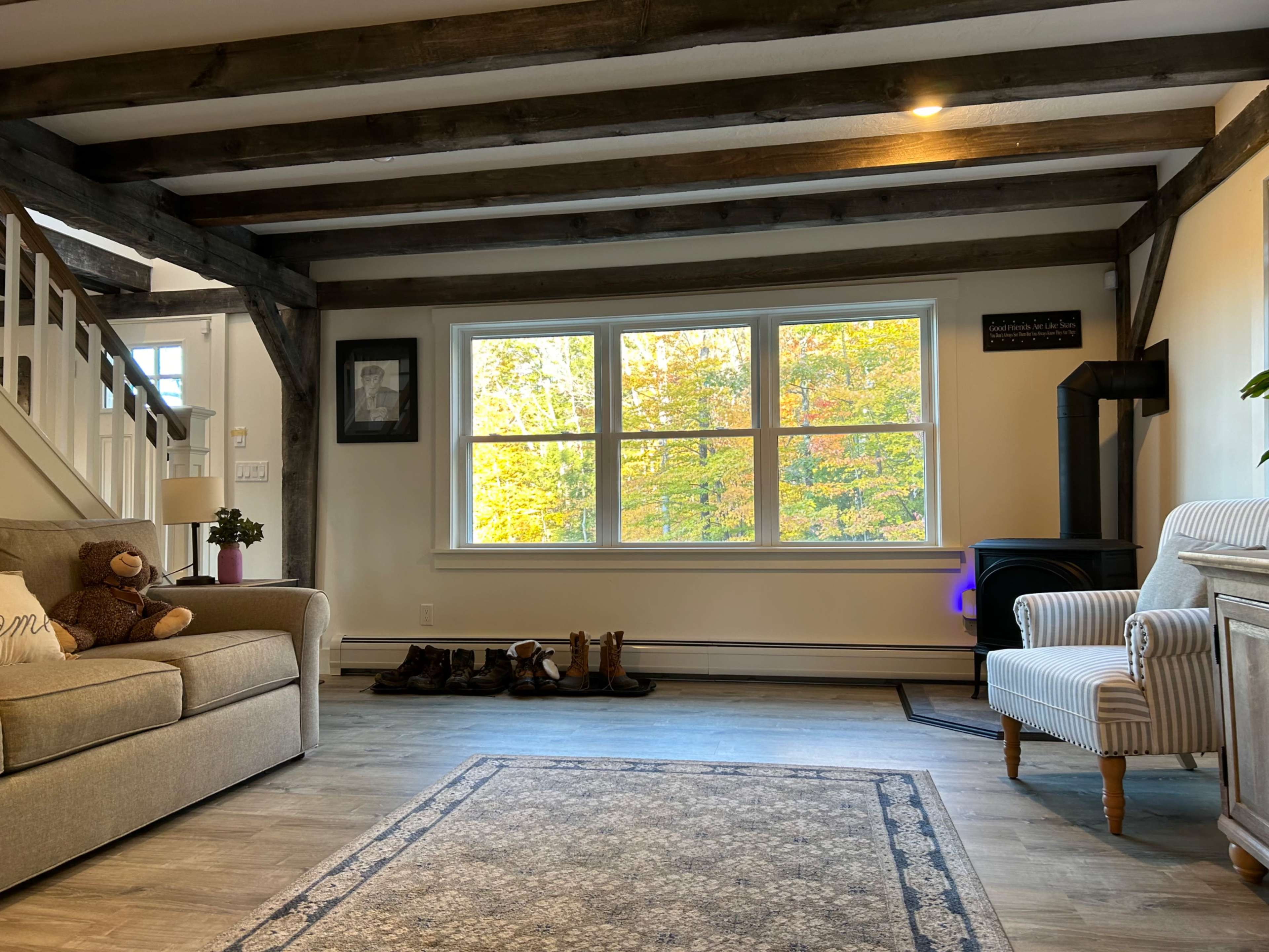 A cozy living room features a sofa and armchair, with a large window displaying colorful autumn foliage outside.