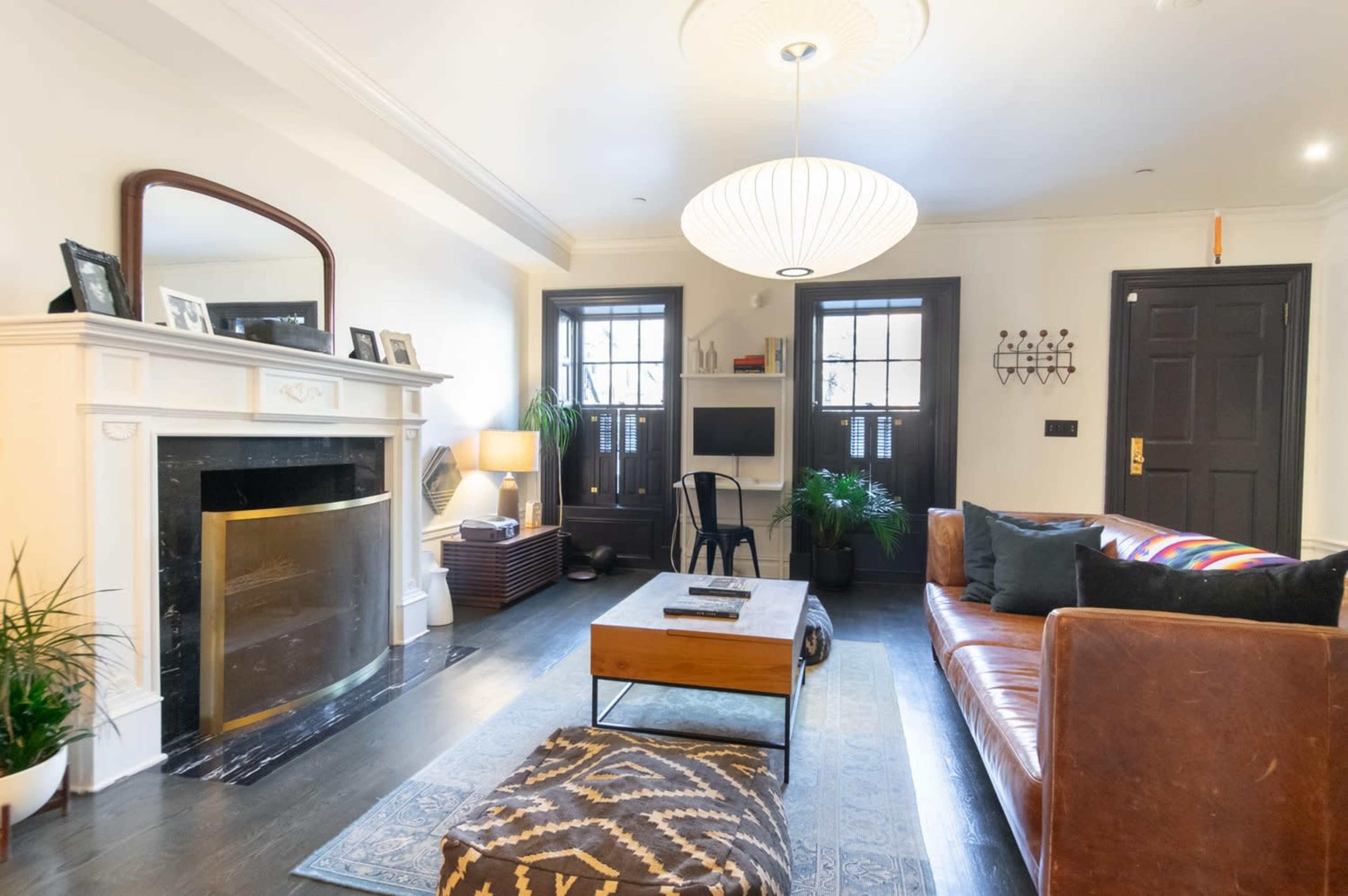The image shows a living room featuring a brown leather sofa, a wooden coffee table, a fireplace with a mirror above it, and two dark doors with windows.